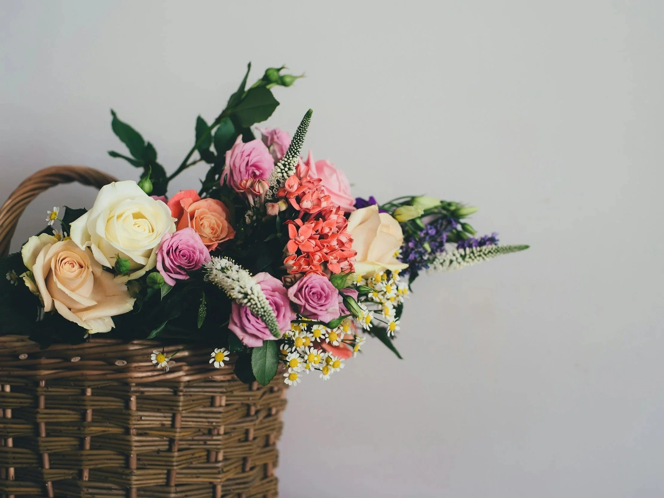 A wicker basket filled with a colorful assortment of fresh flowers, including roses, daisies, and other blooms, placed against a plain gray background.