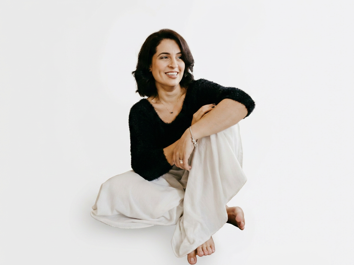 A woman with shoulder-length dark hair sitting on the floor against a plain white background, wearing a black sweater and white pants, smiling while looking to the side.