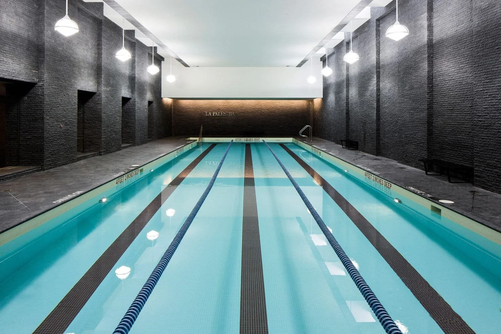 Indoor swimming pool with blue lanes, black walls, and modern light fixtures hanging from the ceiling.