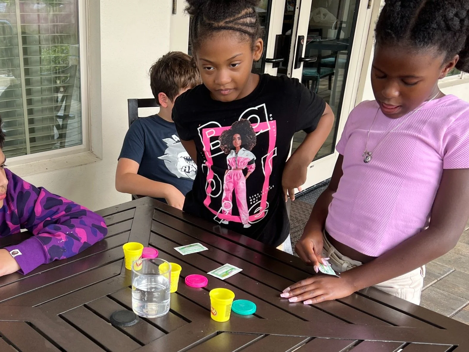 Group of children at a table playing with yellow and pink Play-Doh, clear glass of water, and packets, in an outdoor setting.