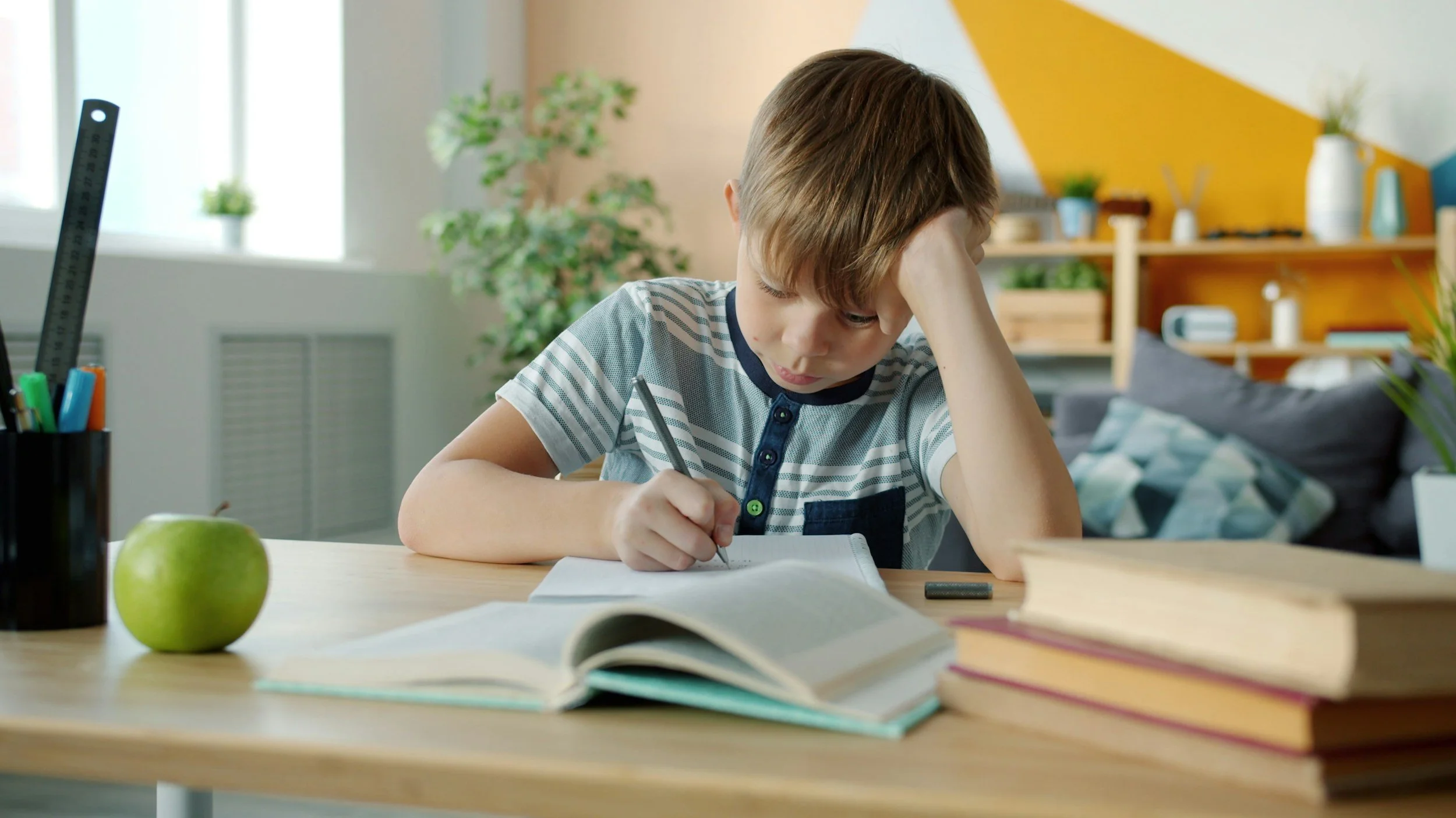 boy in striped shirt sitting at desk writing on paper. Desk has an apple, books and pen holder