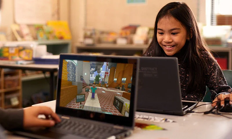 A girl smiling at her laptop, which displays a Minecraft game, in a classroom setting.