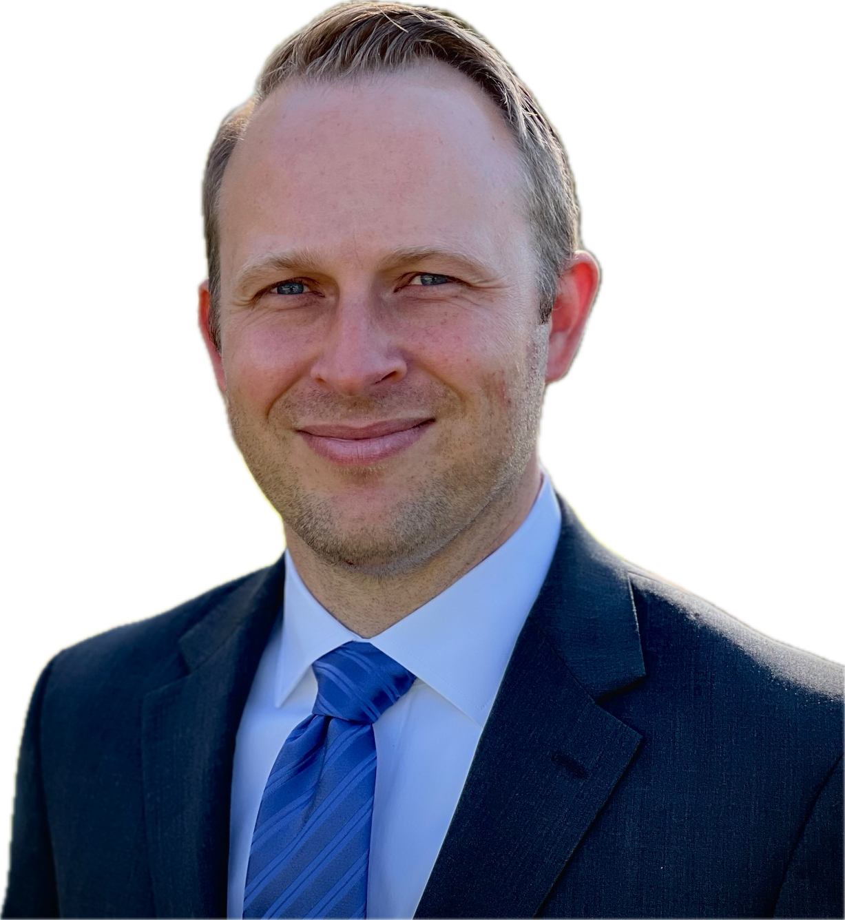 A man with light skin, short light brown hair, blue eyes, wearing a dark suit, blue striped tie, and white shirt, smiling outdoors.