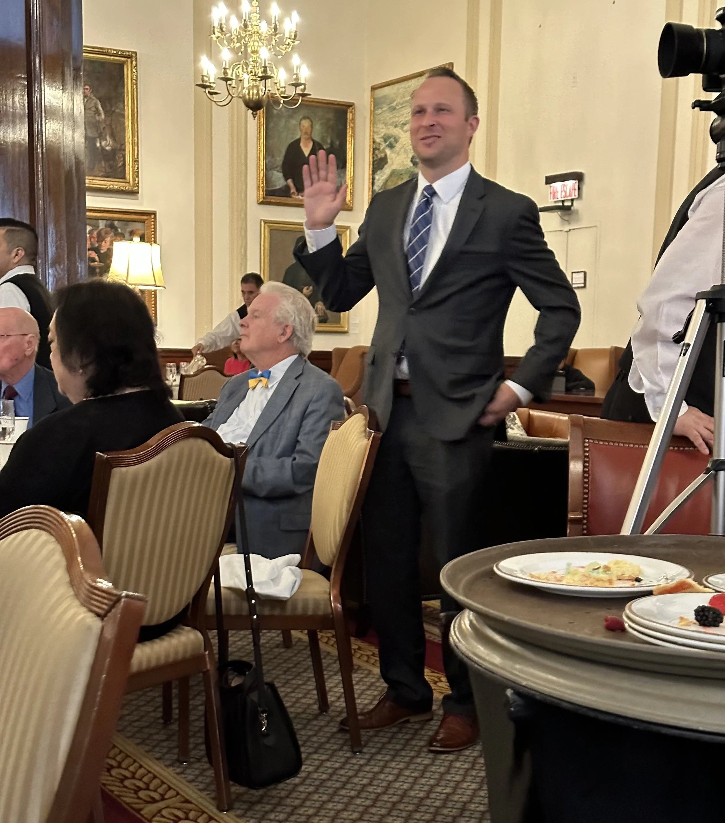 A man in a gray suit standing and raising his right hand in a formal event, with other seated guests around him, in an elegantly decorated room with chandeliers, paintings, and a table with plates of food.