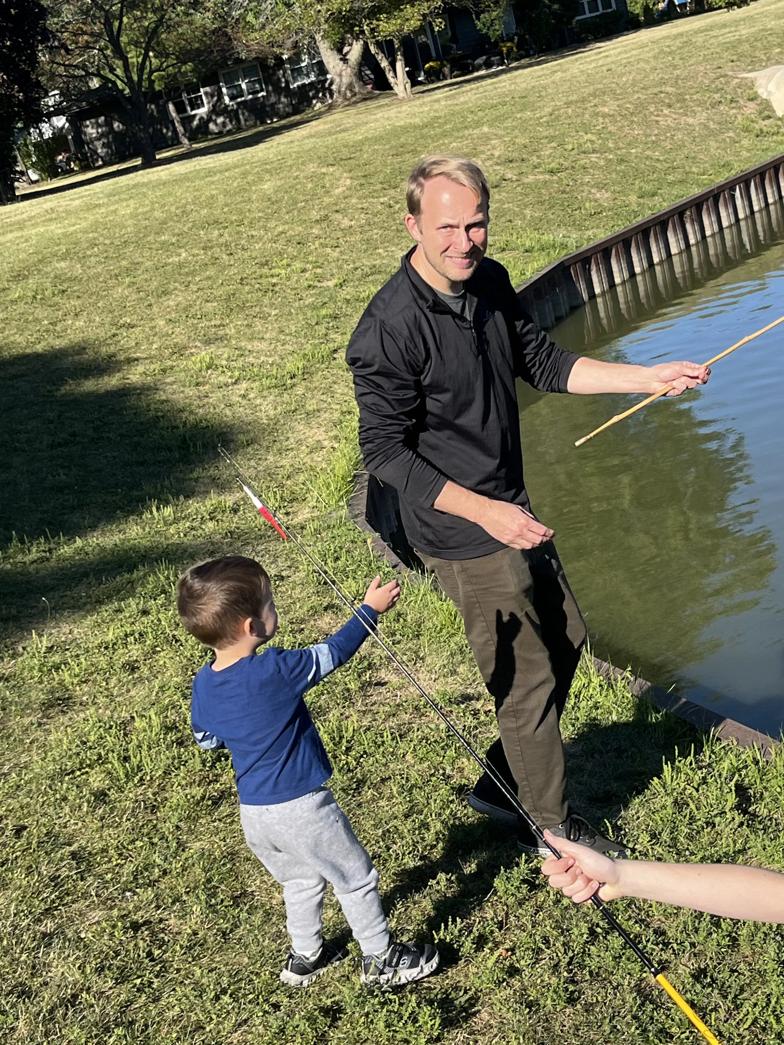 A man and a young boy fishing by a pond on a sunny day in a park.