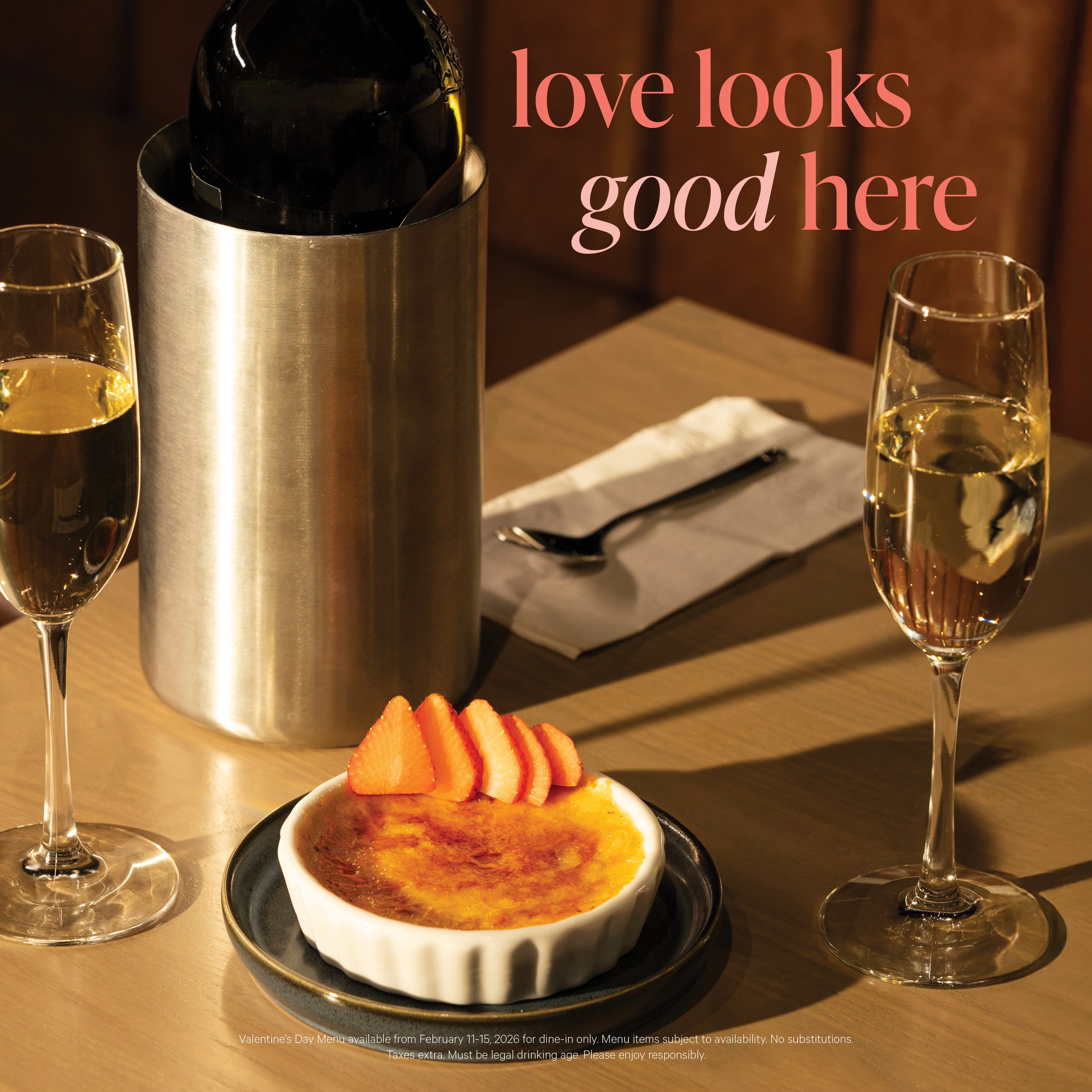 A Valentine's Day dessert with strawberries, a crème brûlée, a bottle of champagne in a silver holder, and two glasses of champagne on a wooden table with a warm background and overlaid text 'love looks good here'.