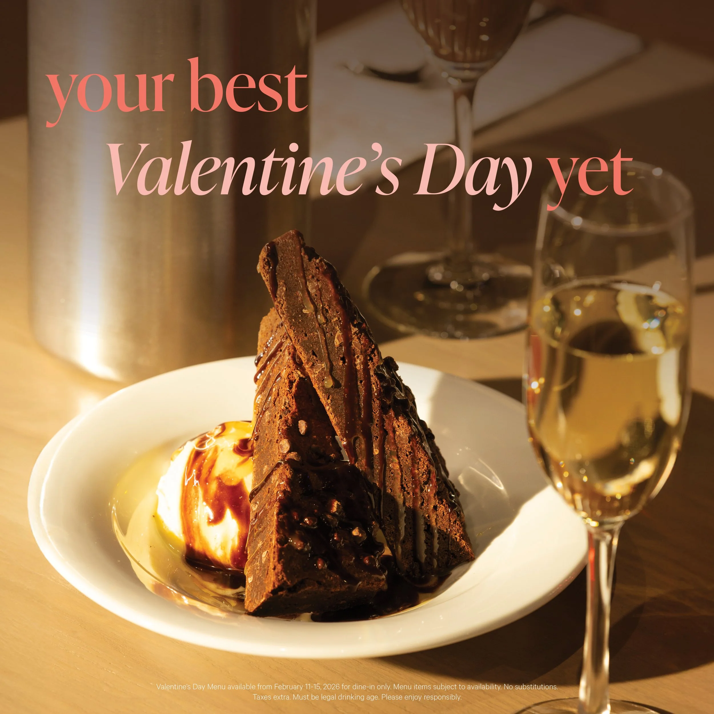 A dessert with a chocolate cake on a white plate, topped with a drizzle of chocolate syrup, accompanied by a scoop of vanilla ice cream with caramel sauce, served alongside a glass of white wine, with a background of a table setup including a metal container, a spoon, and glasses.