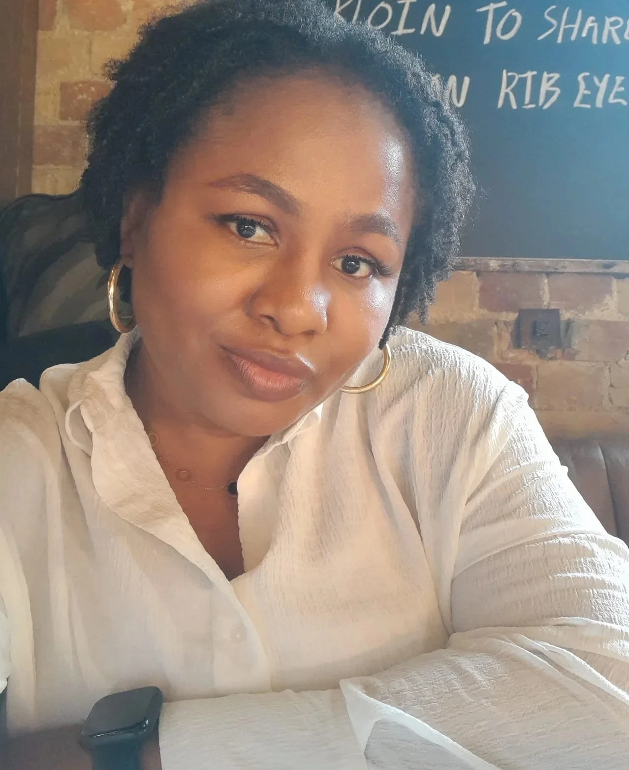 A woman with curly black hair, wearing gold hoop earrings and a white shirt, sitting in a restaurant with a brick wall and a blackboard with white text behind her.