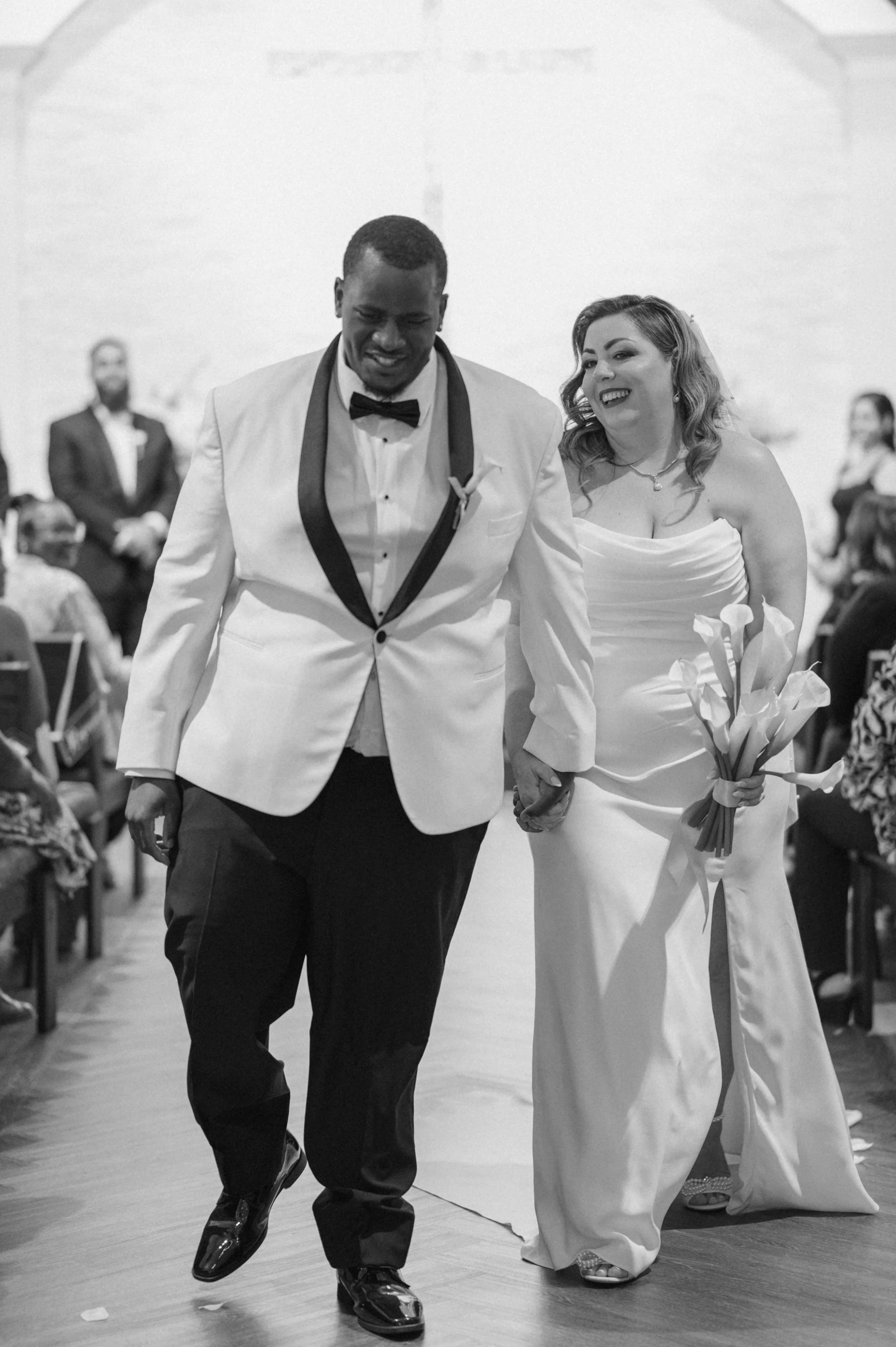 A couple, dressed in wedding attire, walking down the aisle hand in hand at their wedding reception, smiling and happy.