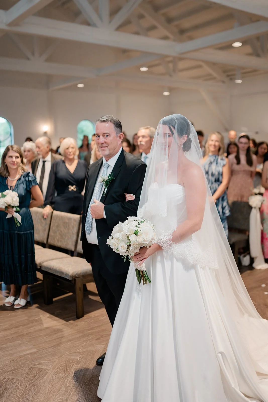 Bride in a white wedding dress with a veil walking down the aisle, holding a bouquet of white roses, accompanied by a man in a black suit, at a wedding ceremony with guests in the background.