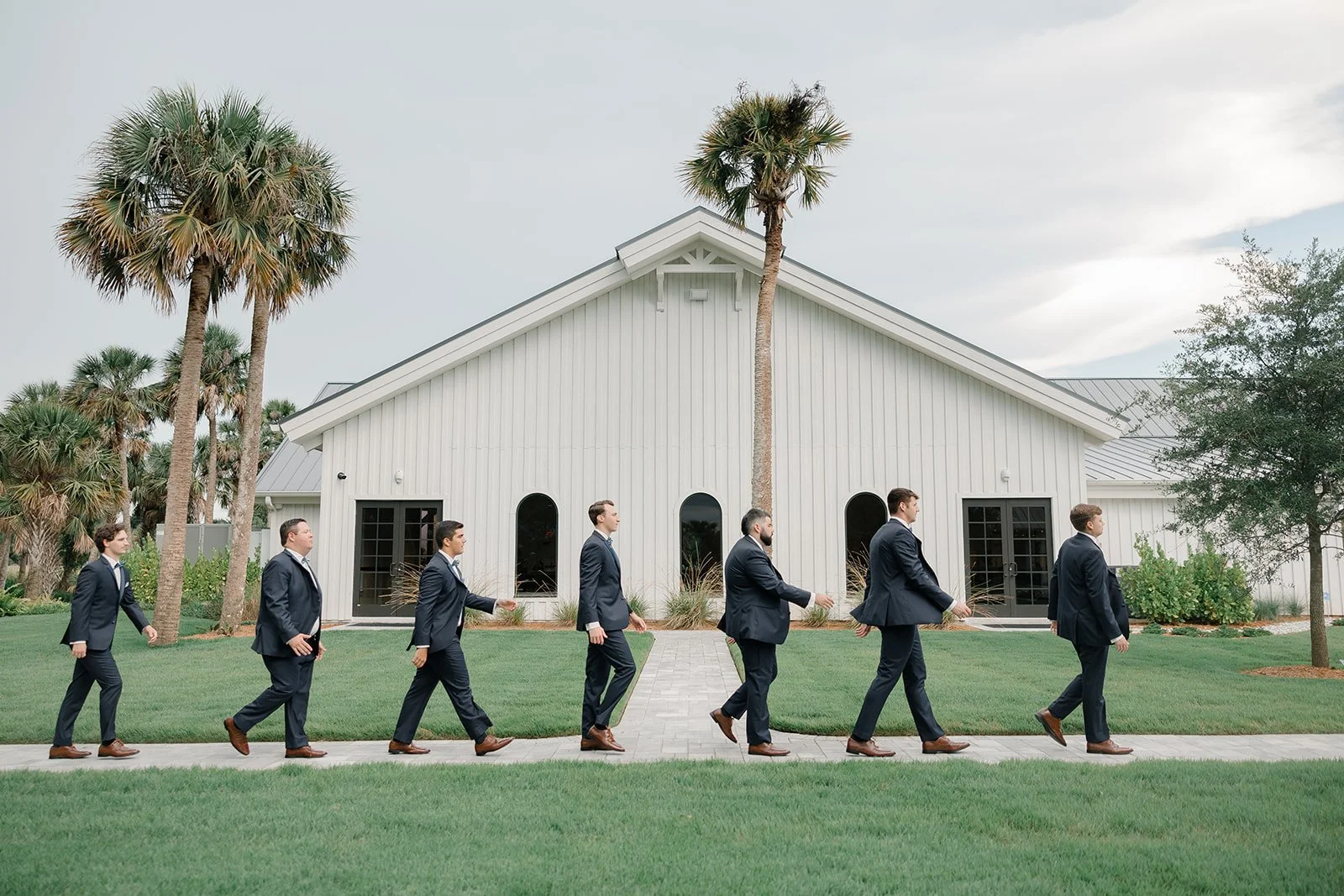 Group of men in suits walking in a line outside a white building with palm trees, on a grassy lawn.