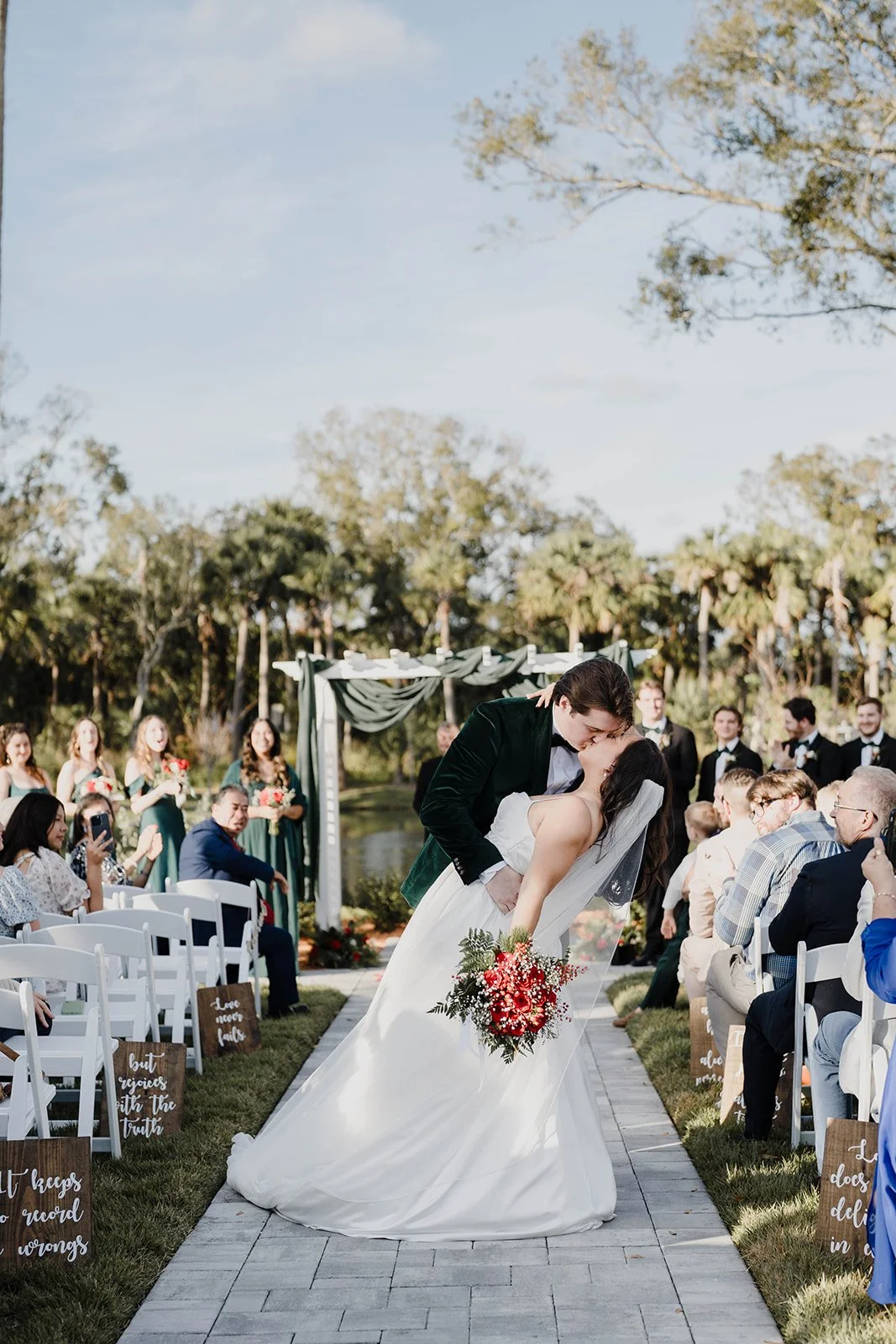 A newlywed couple sharing a kiss while dancing outdoors during their wedding ceremony. The bride is holding a bouquet of red and pink flowers, and guests are seated on white chairs on either side of the aisle with signs. The scene is set in a lush ga
