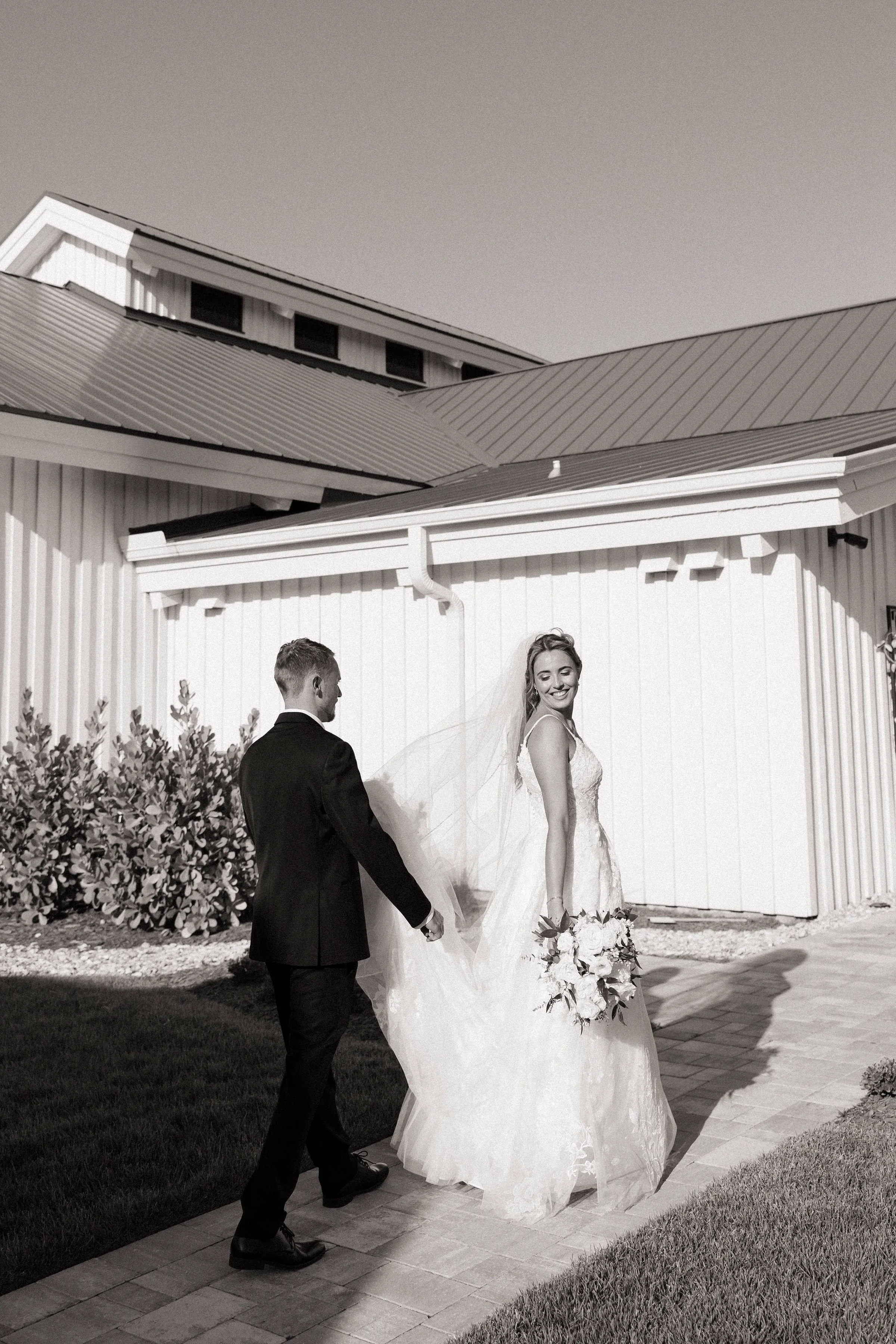 A black-and-white photo of a bride in a wedding dress holding a bouquet of flowers smiling while walking outside with a groom in a suit during daytime.