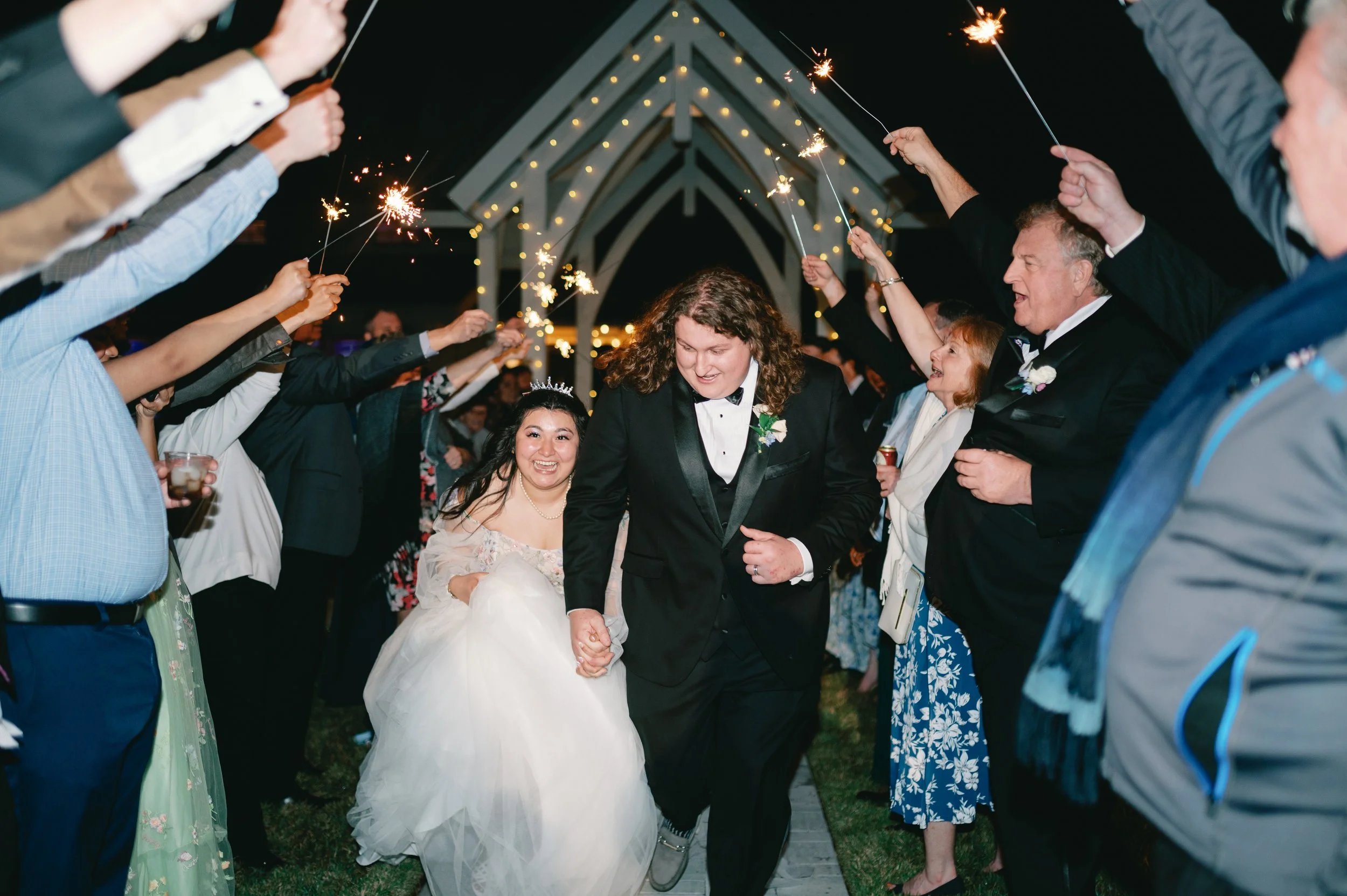 Couple, one in a tuxedo and the other in a wedding gown, holding hands and walking through a tunnel of sparklers held by friends and family during a wedding celebration at night.