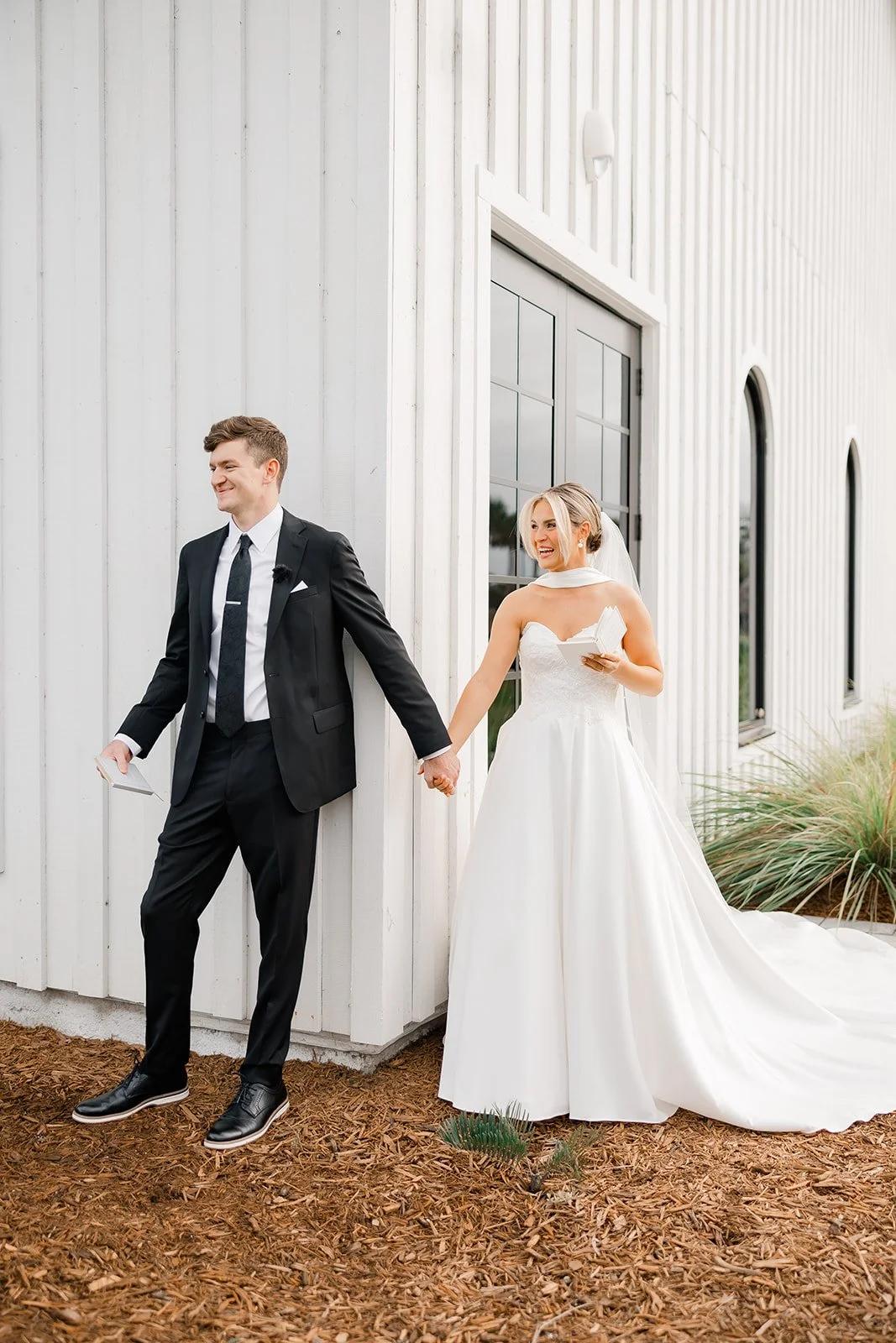 A bride and groom stand outside a white building, holding hands and smiling, during their wedding. The bride is in a white wedding dress, and the groom is in a black suit.
