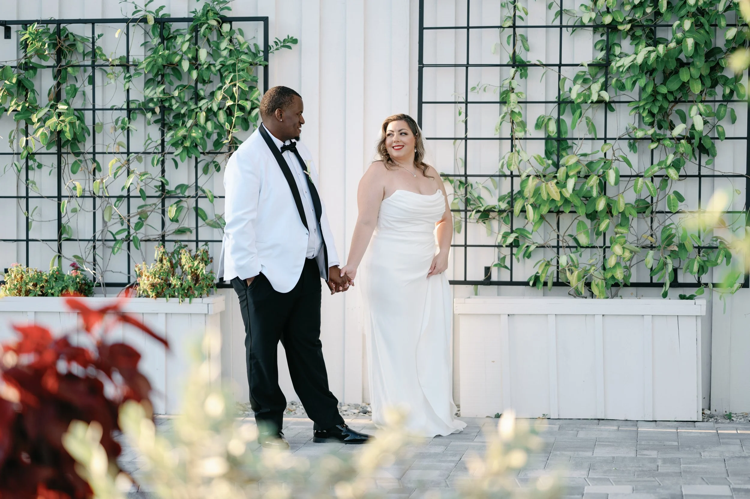 A bride and groom holding hands and looking at each other outdoors. The bride is wearing a white strapless wedding gown, and the groom is dressed in a white tuxedo jacket with black pants and a black bow tie. They are standing in front of a white fen