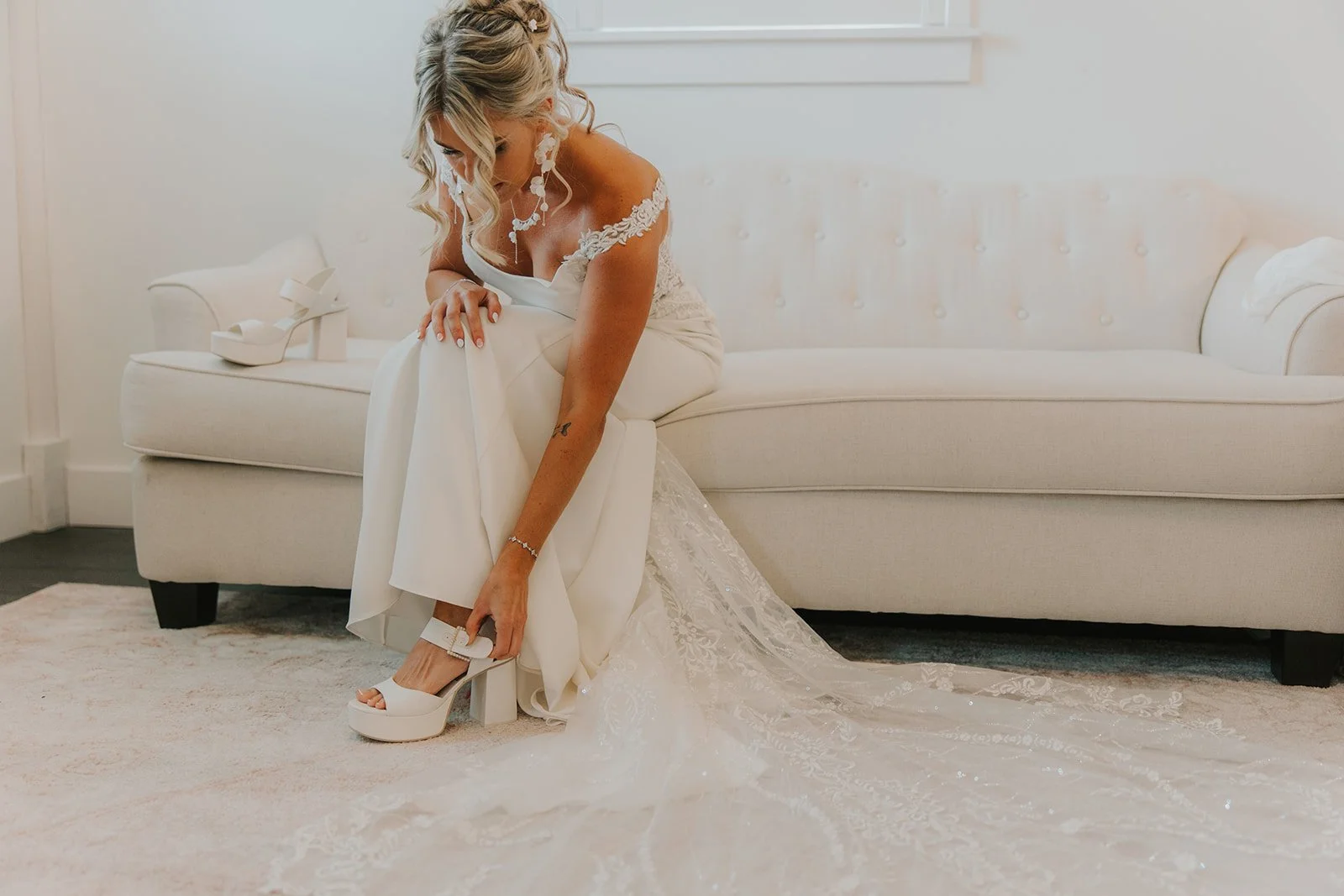 Brunette bride in an elegant white wedding dress getting ready, sitting on a beige sofa, putting on white high-heeled shoes with ankle straps, with her wedding gown's long lace train spread on the floor beside her.
