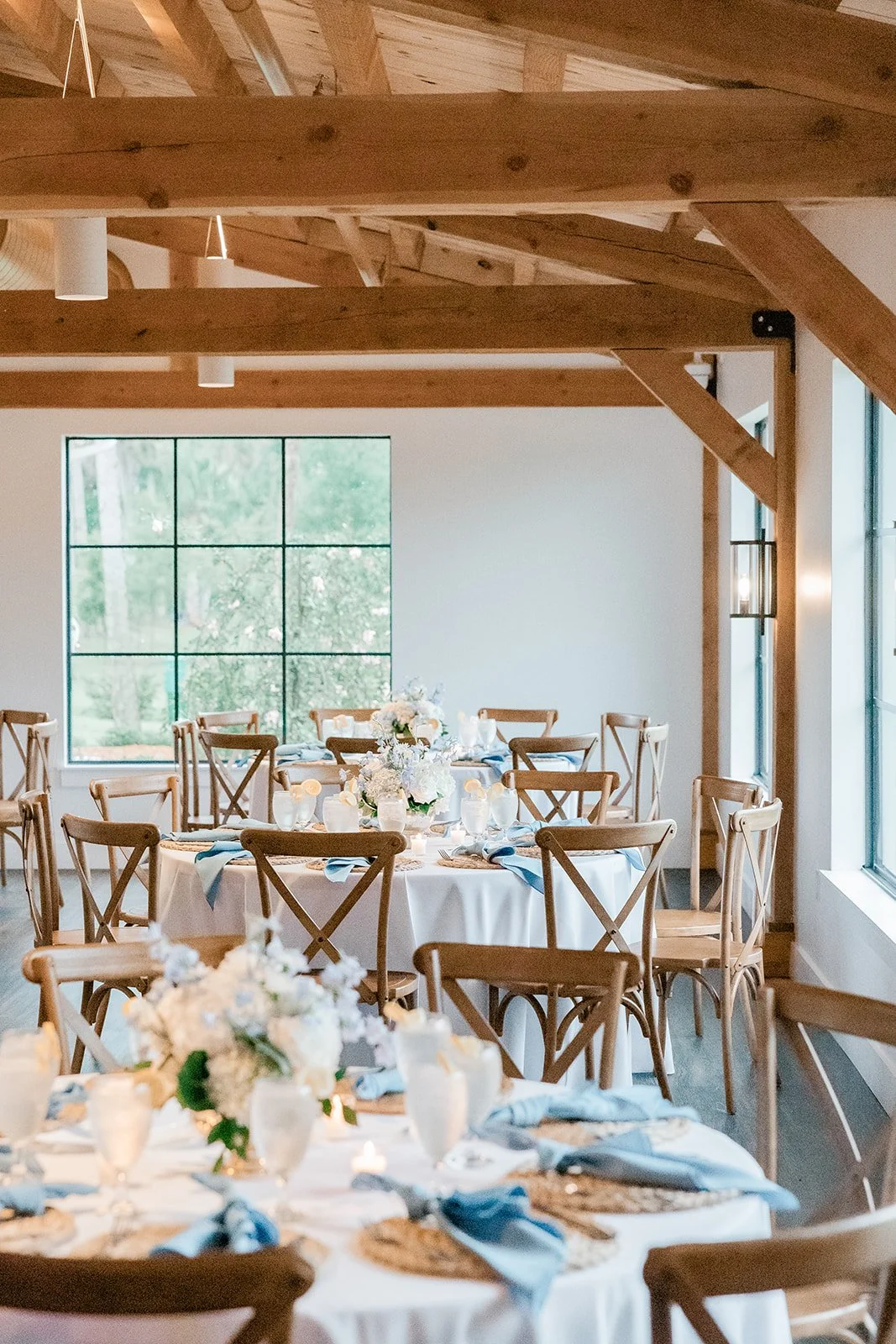 Indoor dining area with round tables decorated with white tablecloths, flower centerpieces, and blue napkins, surrounded by wooden chairs. Large windows let in natural light, revealing green outdoor scenery.