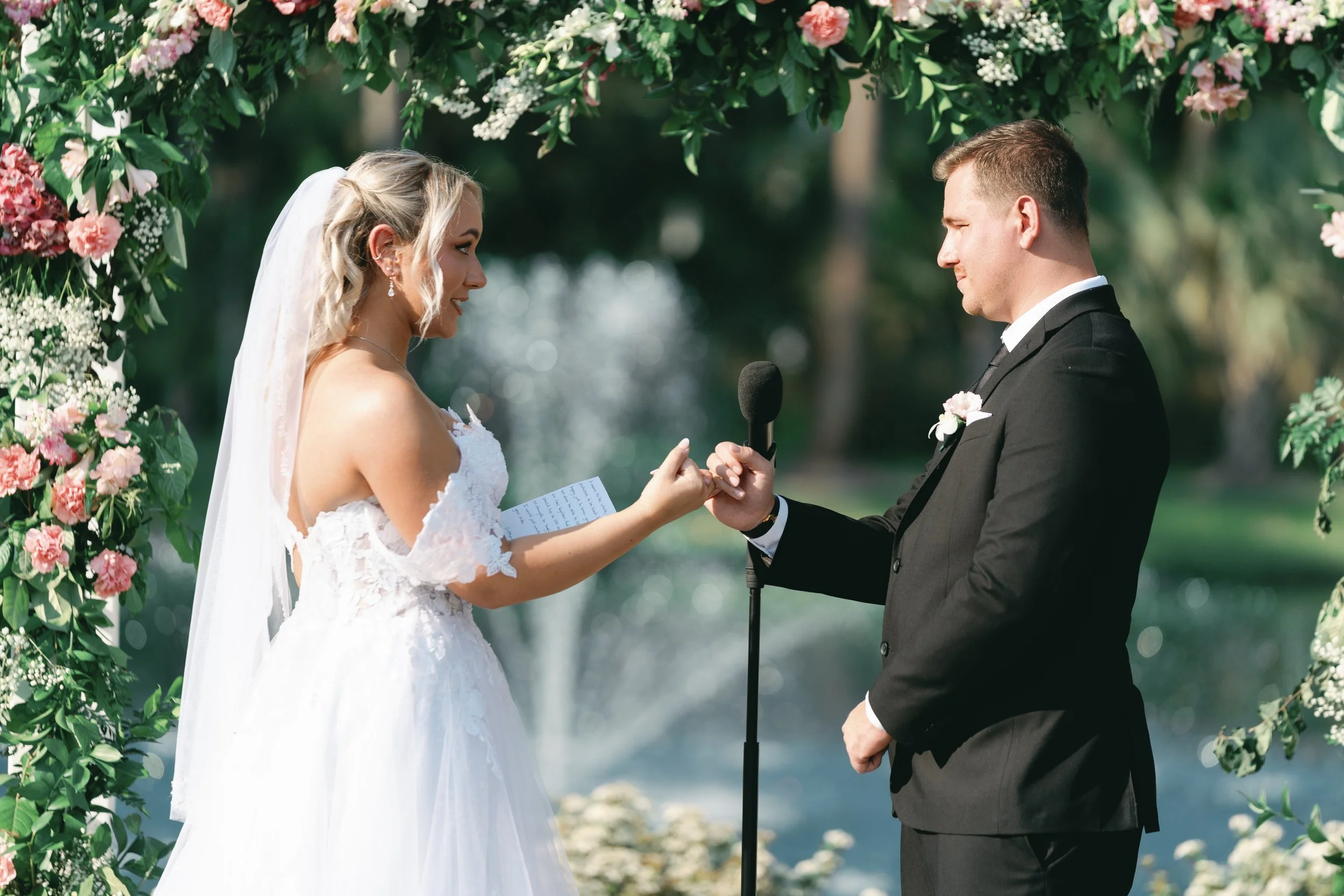 A bride and groom exchanging wedding vows during an outdoor ceremony under a floral arch, with water and trees in the background.