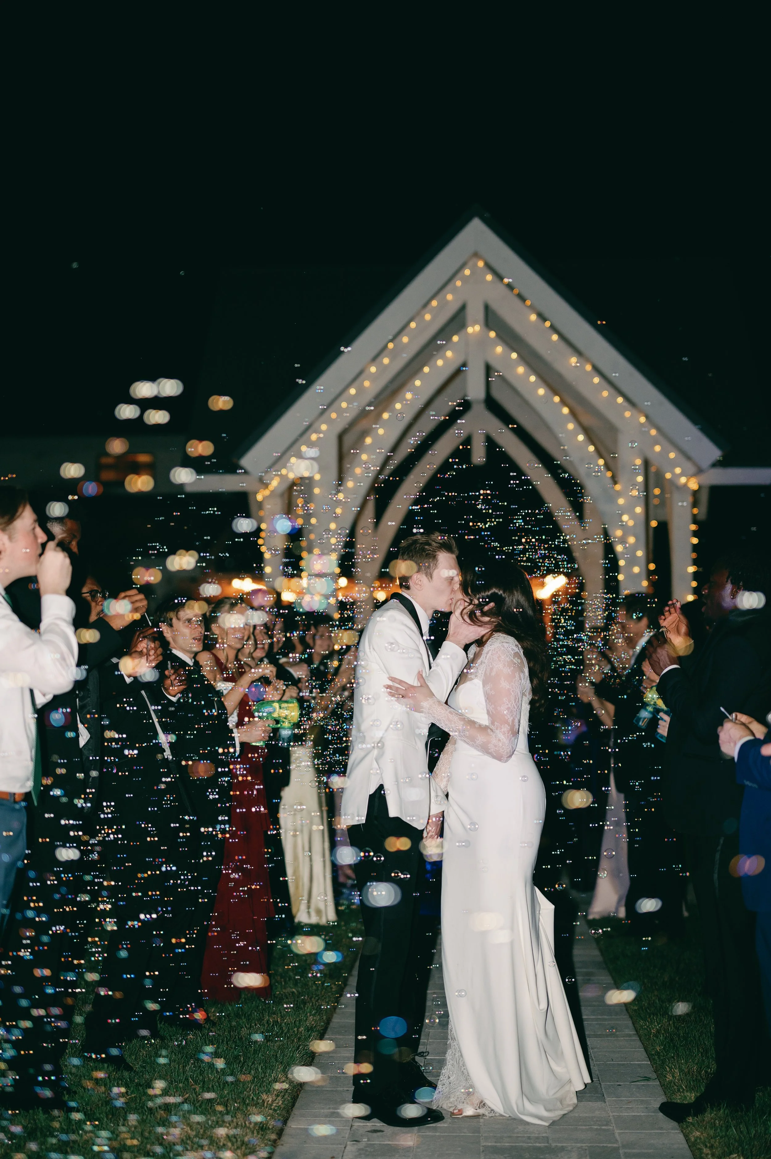 A bride and groom kiss at their wedding ceremony during the night, surrounded by friends and family, with festive lights and a white pergola in the background.