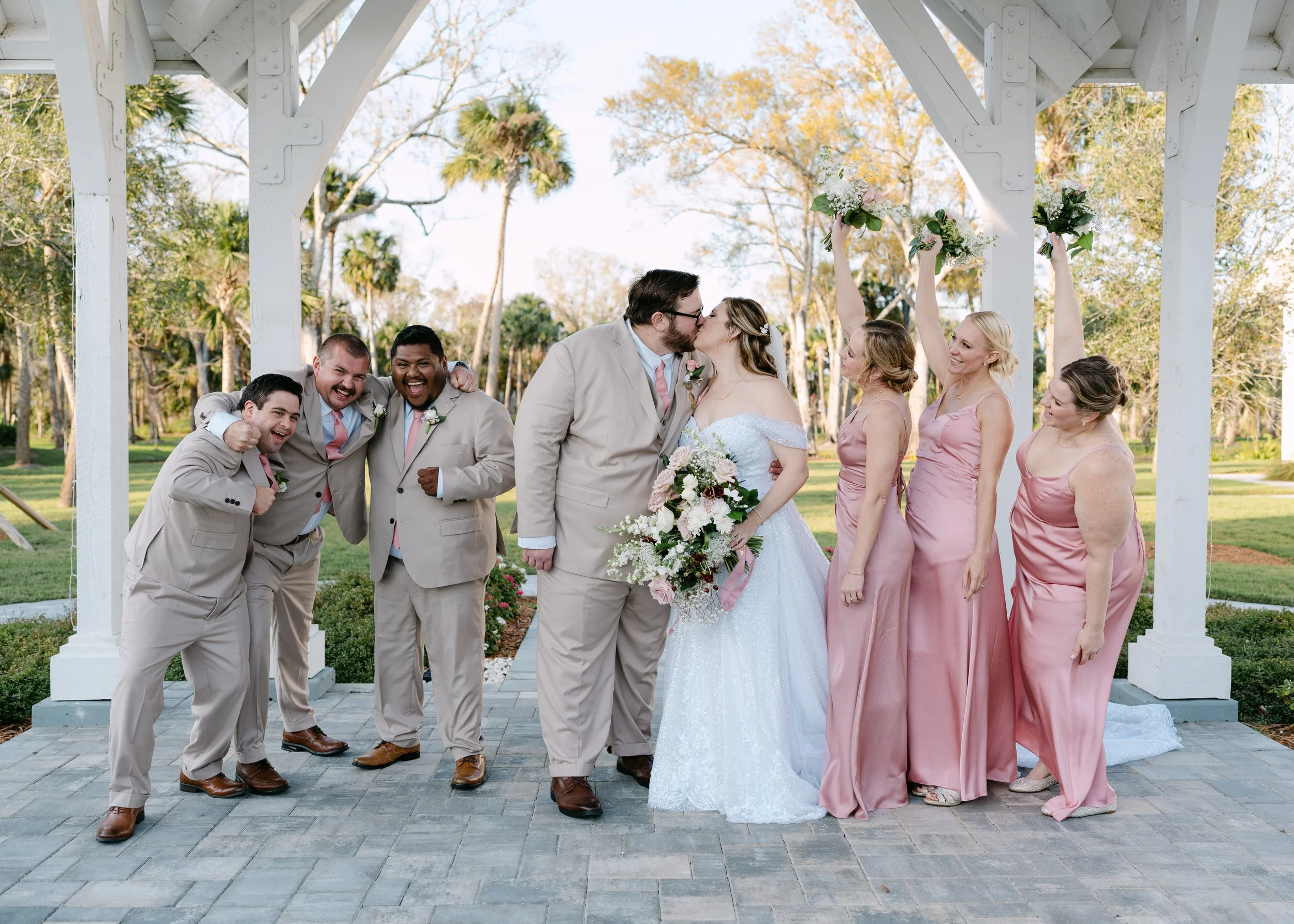 A wedding party under a white gazebo outdoors, with the bride and groom kissing in the center, surrounded by bridesmaids holding bouquets up, and groomsmen celebrating.