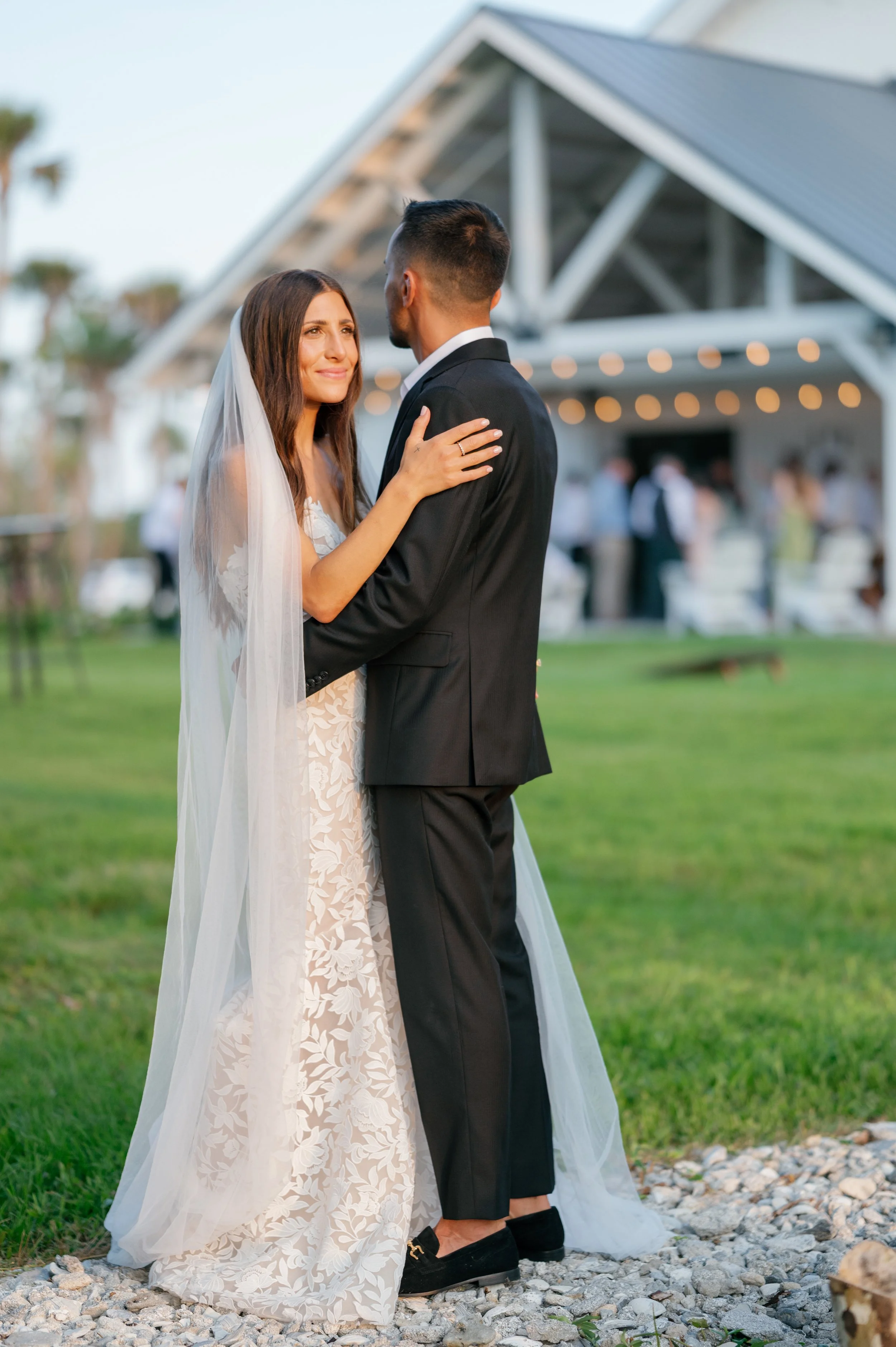 A bride and groom sharing a dance outdoors at a wedding reception, with a barn and guests in the background.