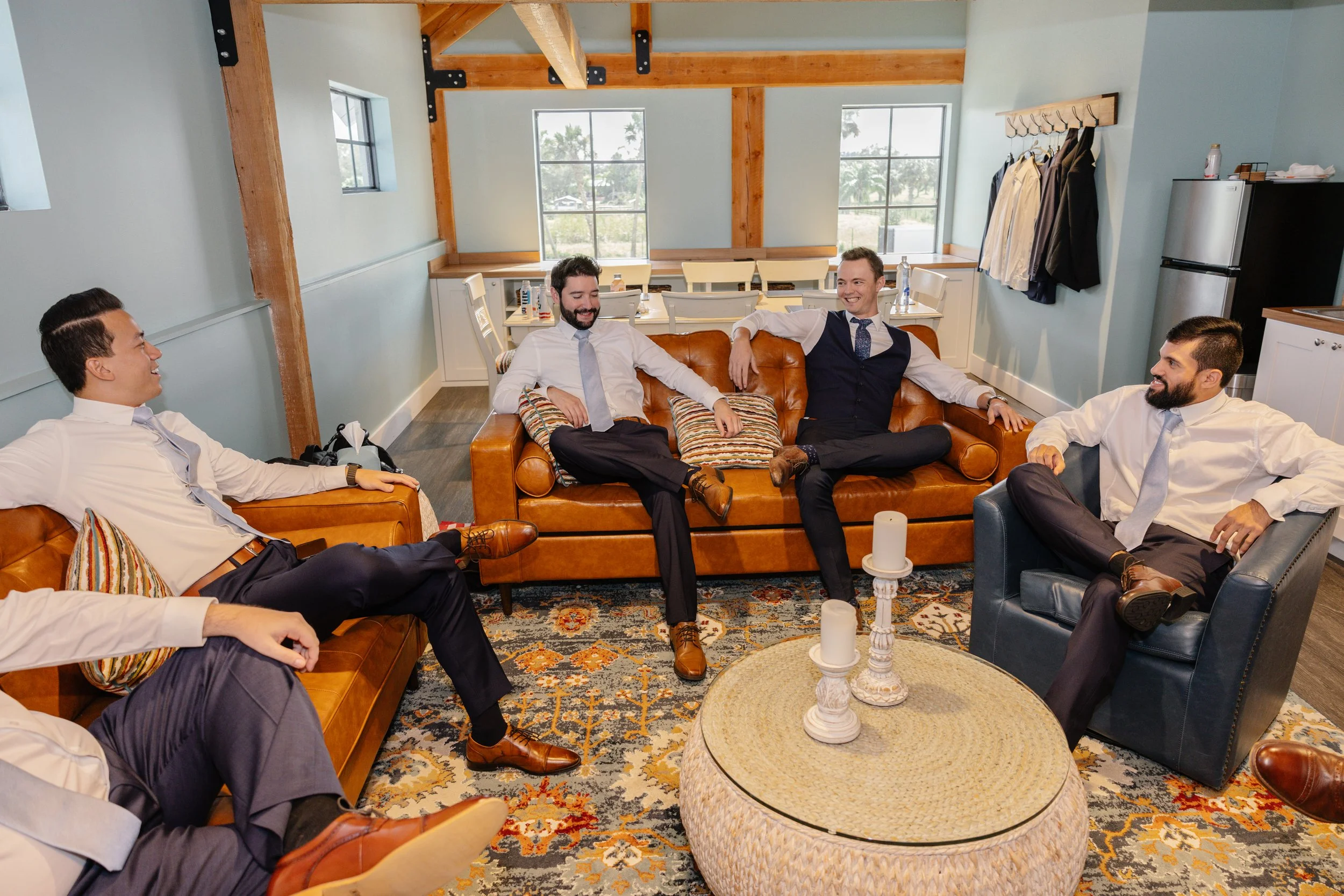 Five men dressed in formal attire are sitting and chatting in a cozy, modern living room with light blue walls, wooden beams, and large windows.