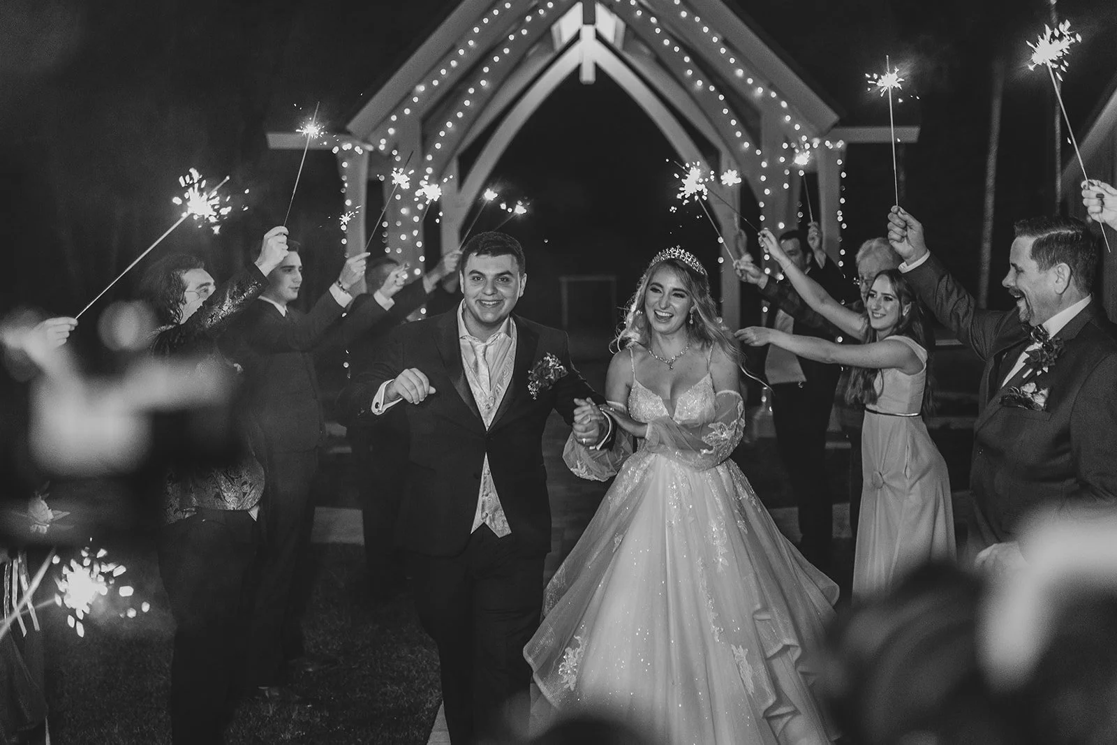 A bride and groom walking hand-in-hand through a celebration with guests holding sparklers at night.