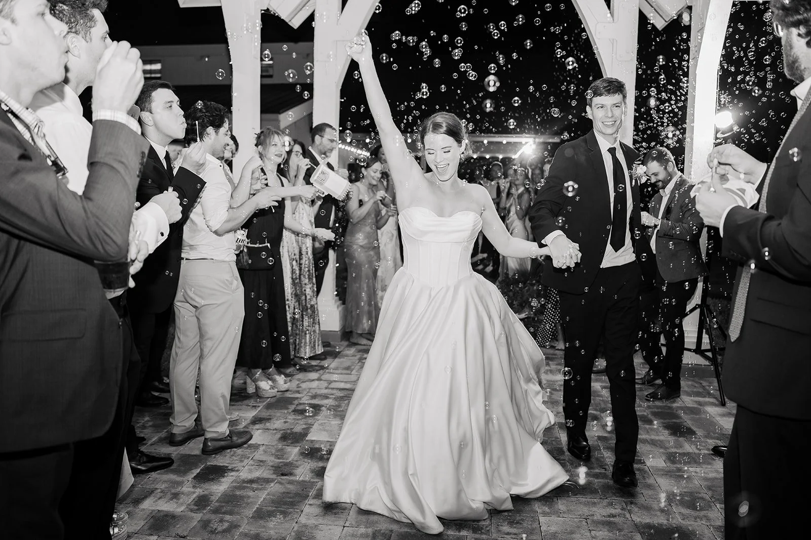 A newlywed couple dance at their wedding reception, surrounded by guests, with bubbles floating around in a decorated venue.