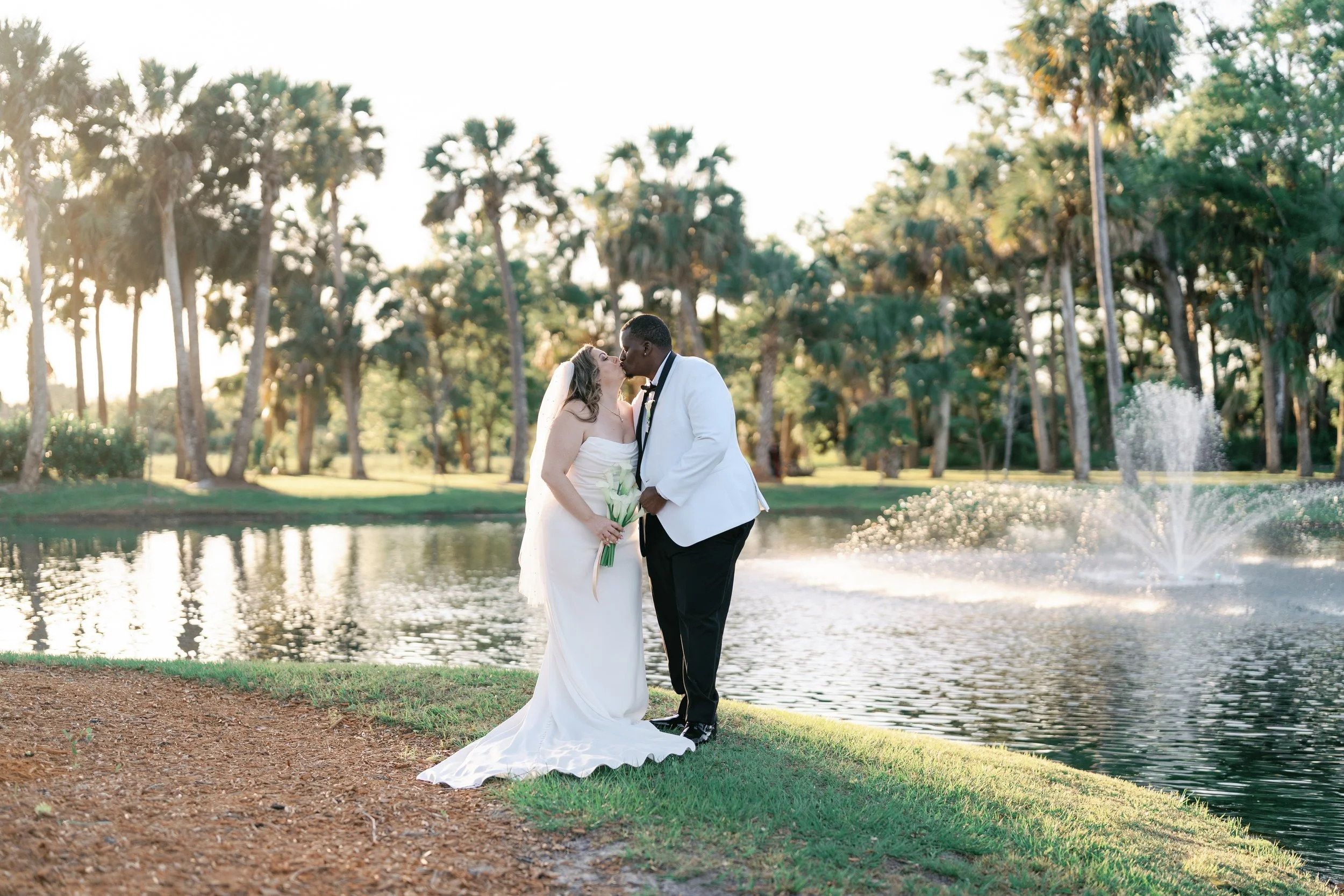 Bride and groom sharing a kiss by a pond during sunset, wedding attire, trees and fountain in background.