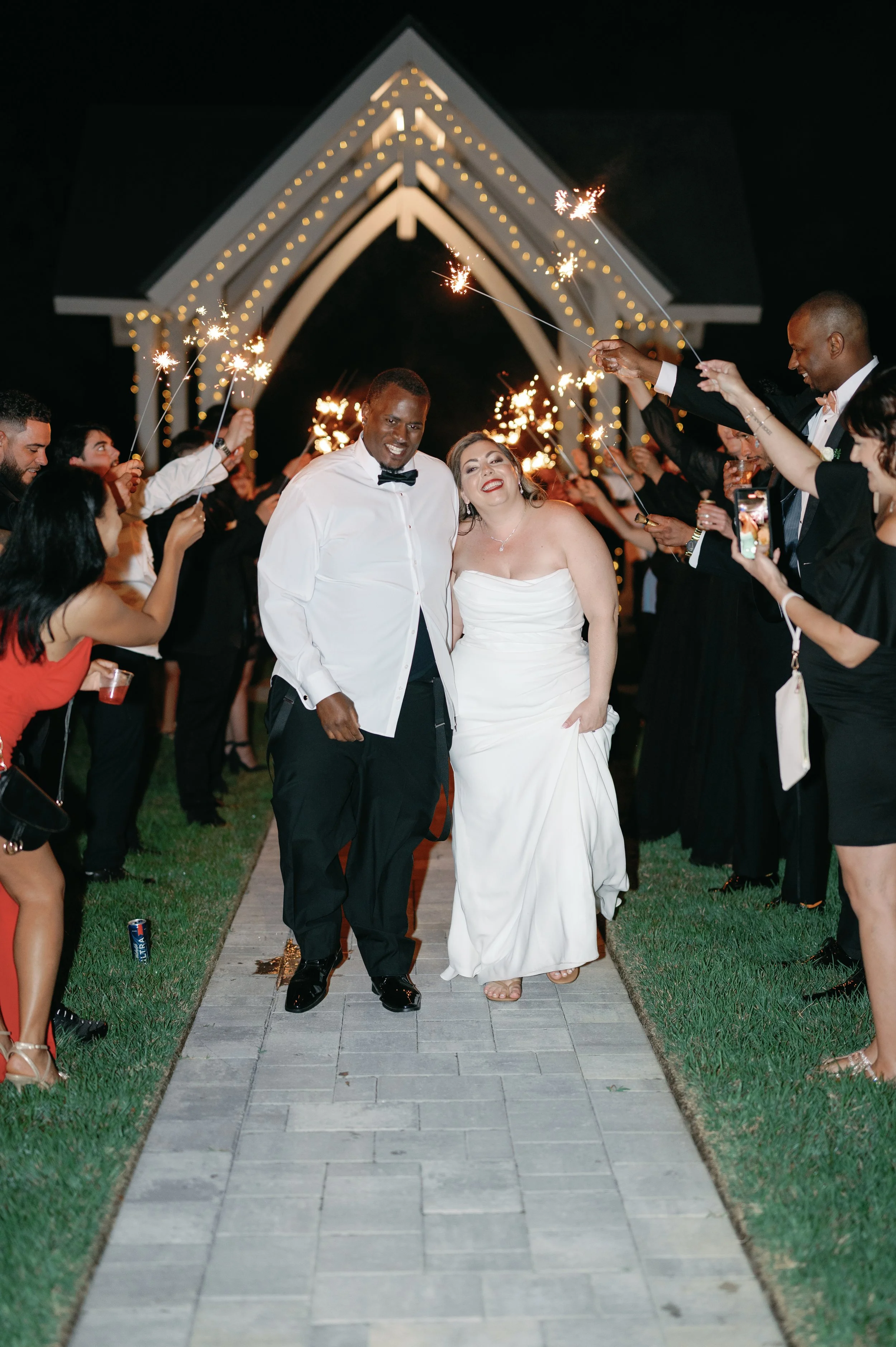 Couple in wedding attire walking down aisle decorated with sparklers and lights at night, celebrating wedding reception.