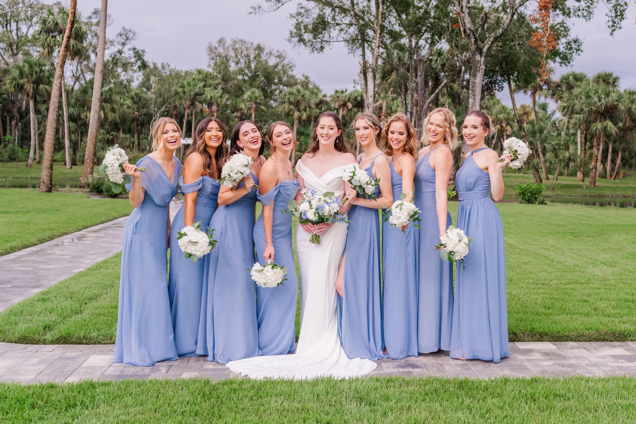 A group of nine women in light blue and white dresses standing on a stone pathway in a park with green grass and trees, smiling and holding bouquets of white flowers, celebrating a wedding.