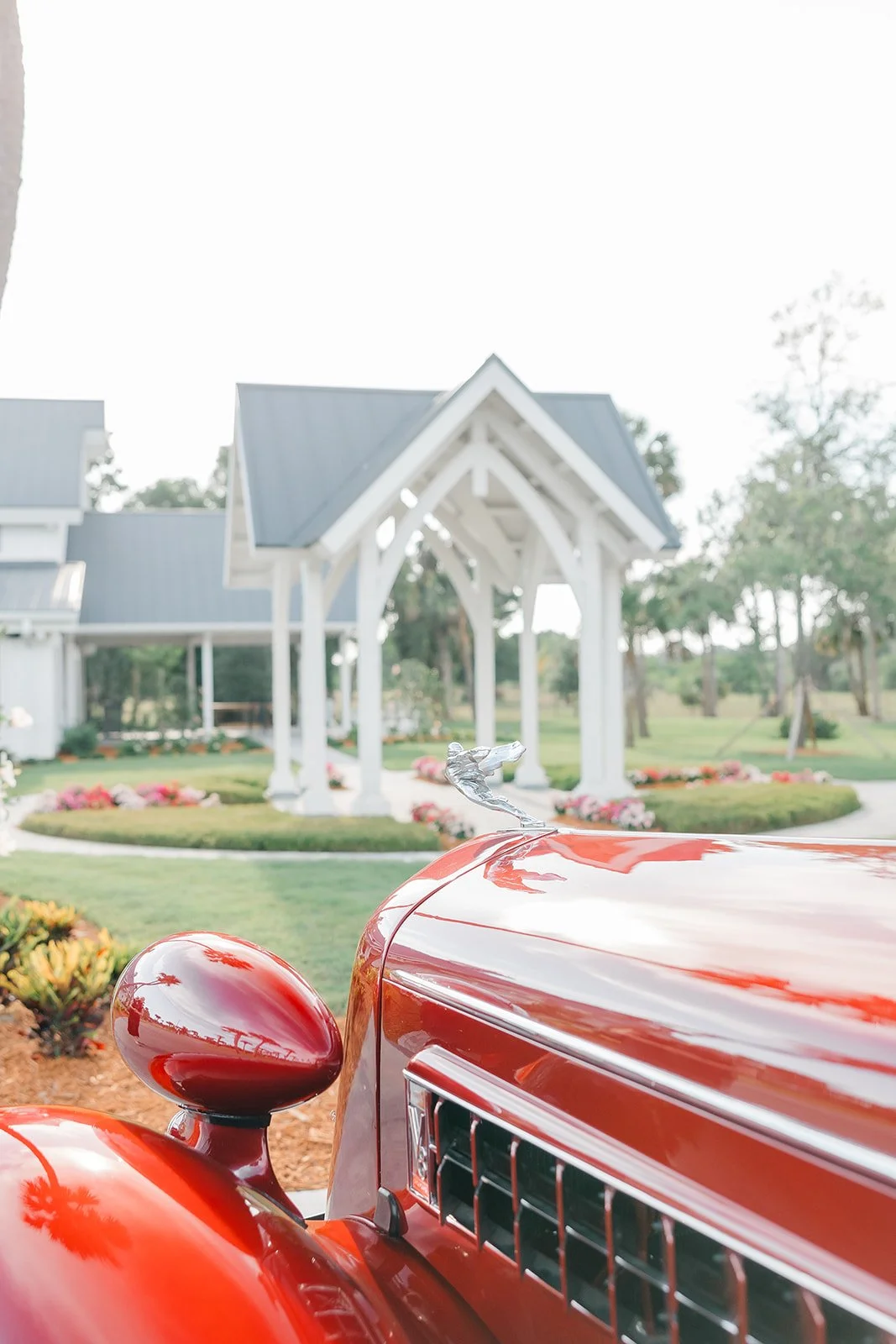 Close-up of a shiny red vintage car hood ornament in the shape of a bird, with a white wooden church and a landscaped garden with flowers in the background.