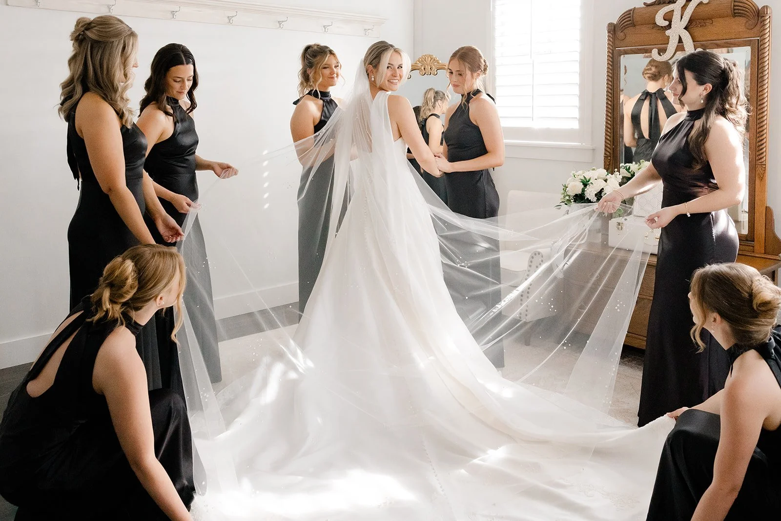 A bride in a white wedding dress, smiling, surrounded by six bridesmaids in black dresses, helping her with her veil in a well-lit room with white walls and a wooden dresser with a mirror.