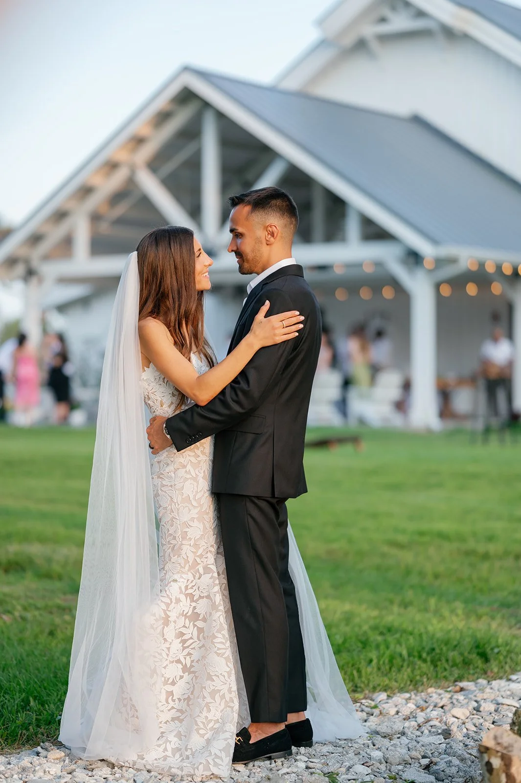 A bride and groom embrace during their outdoor wedding at Celebration Grove Wedding and Event Venue, with a white pavilion and guests in the background.
