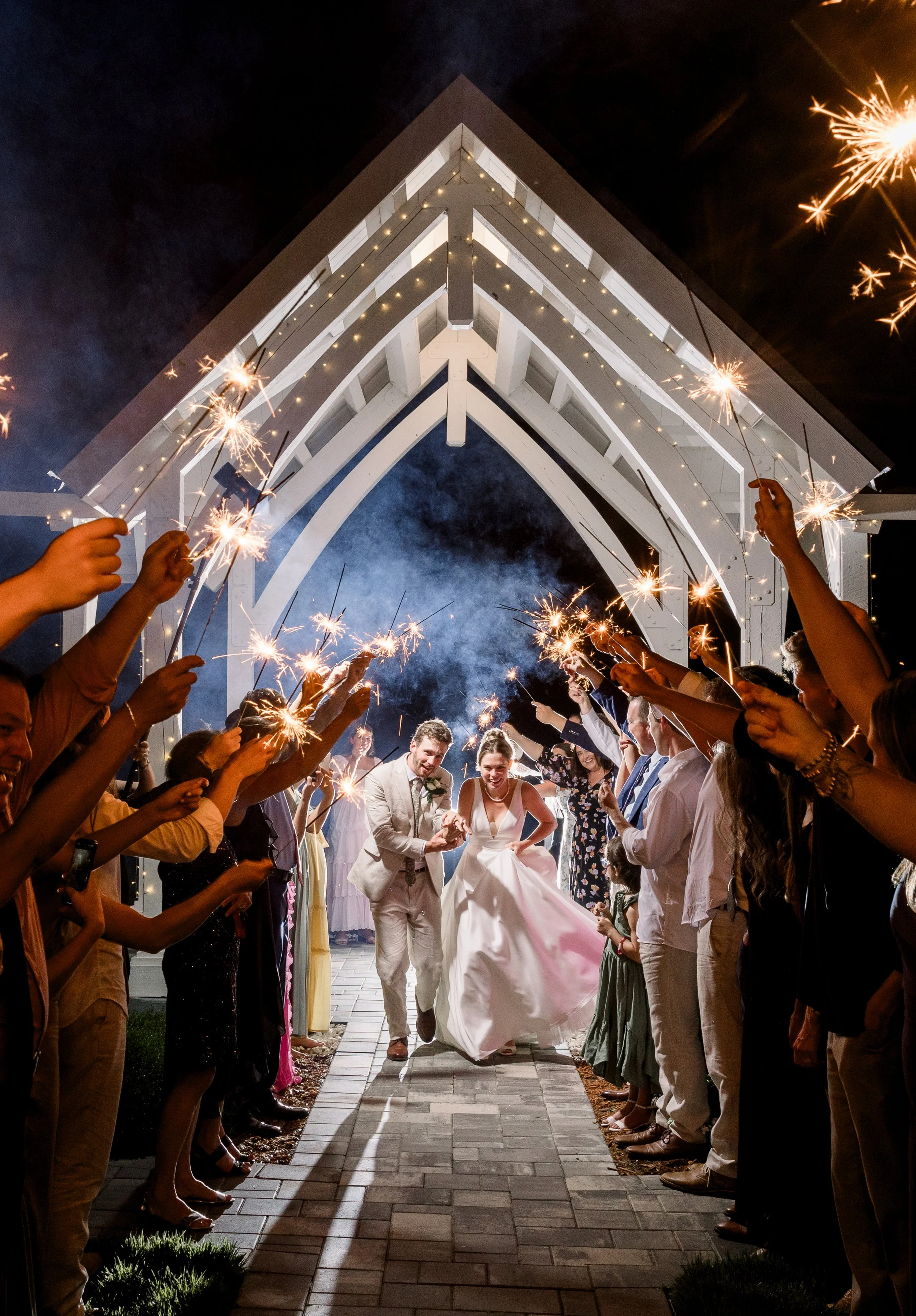 Wedding celebration at night with bride and groom walking under an illuminated archway, holding sparklers during their send-off after their wedding at Celebration Grove, surrounded by friends and family in Vero Beach.