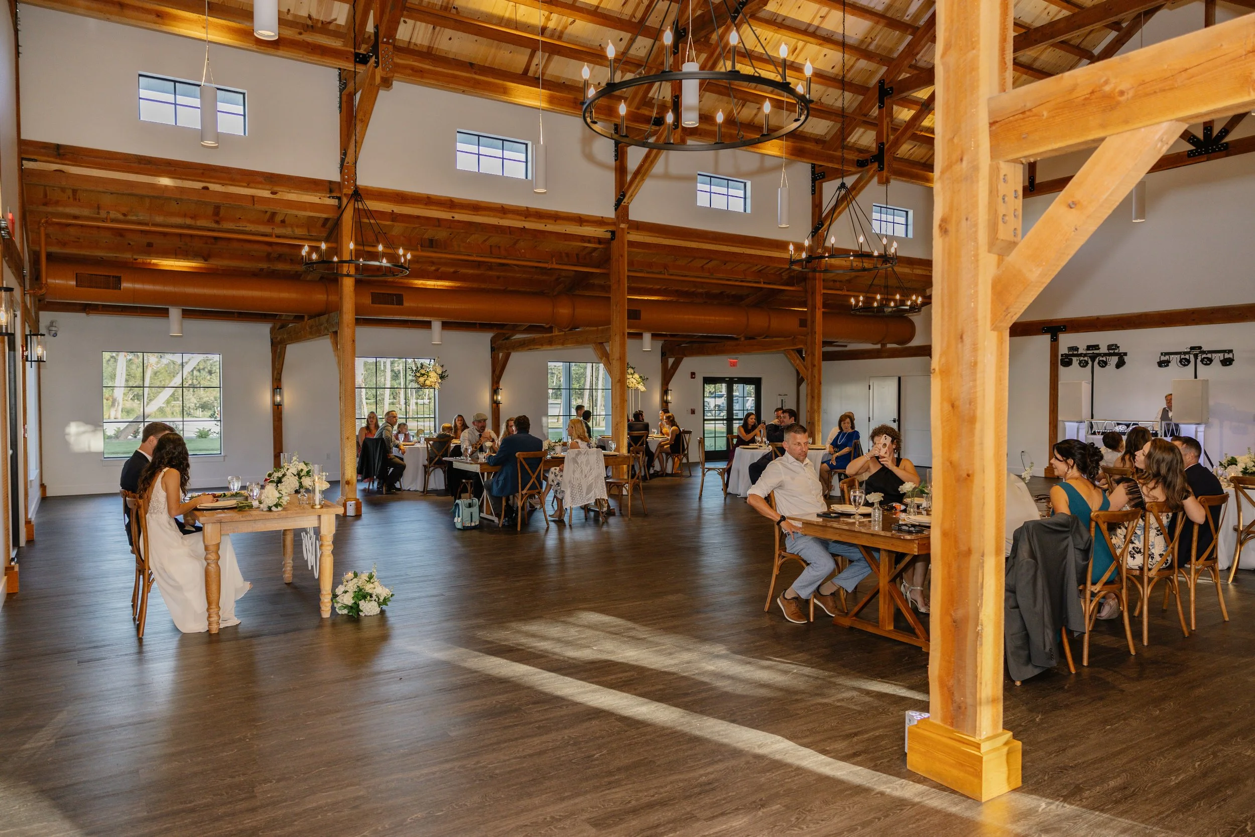 Wedding reception in a rustic, wooden hall with guests seated at tables, chandeliers hanging from the ceiling, and natural light coming through windows.