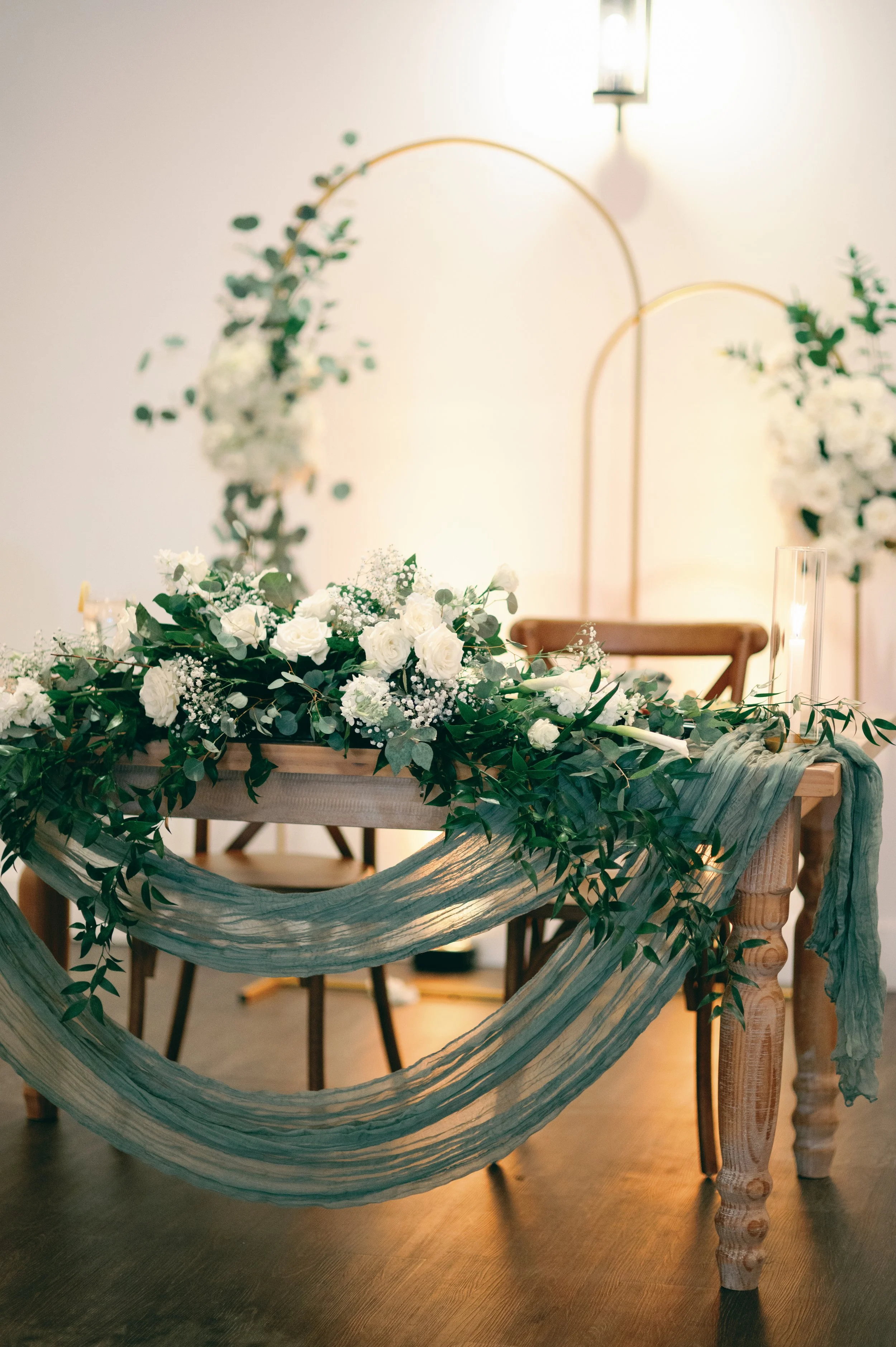 A decorated wooden table with floral arrangements, green drapery, and candles, set for a wedding or special event with a minimalist background.