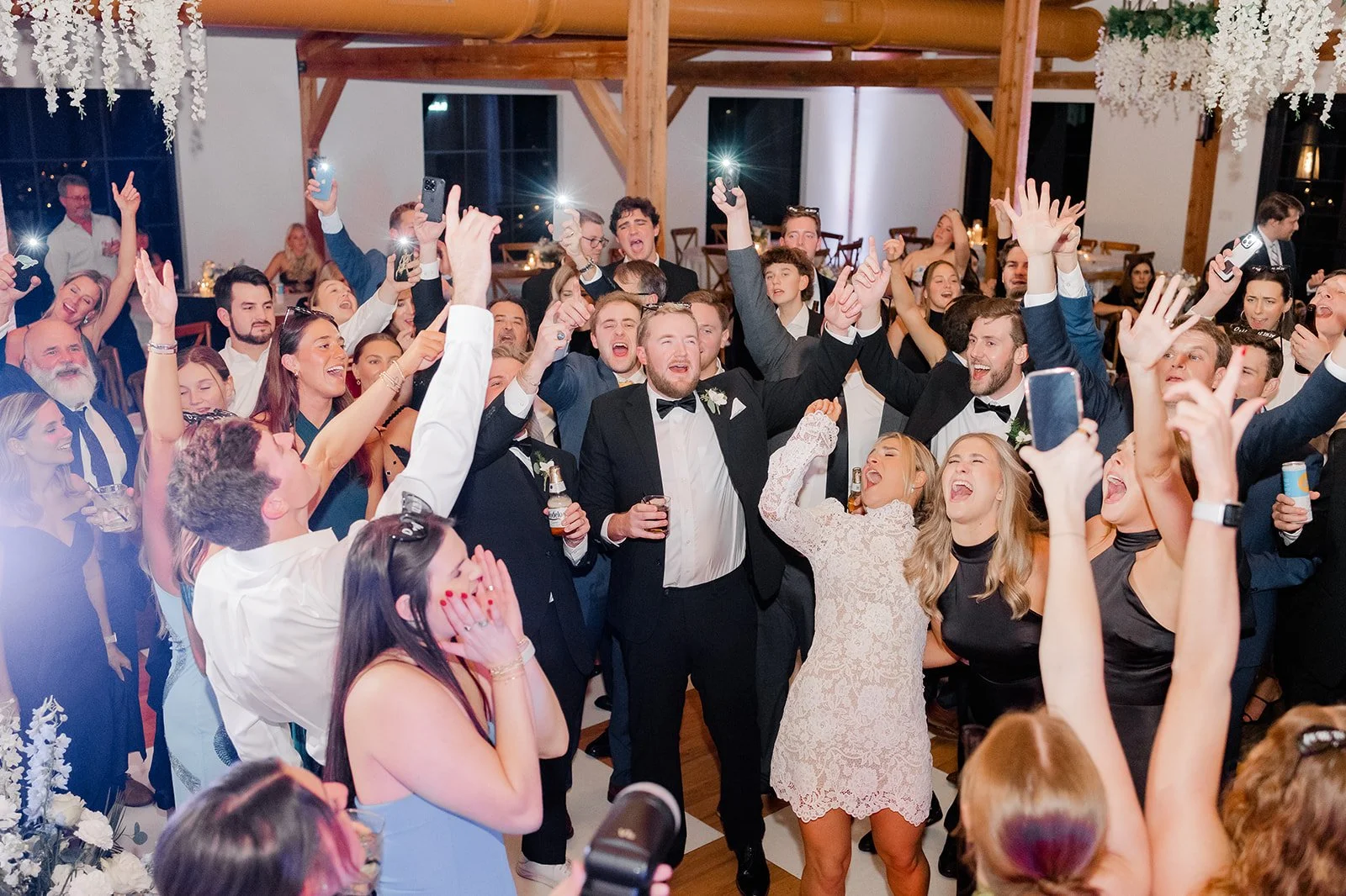 A group of people at a wedding celebration, dressed in formal attire, dancing, singing, and taking photos in a decorated venue.