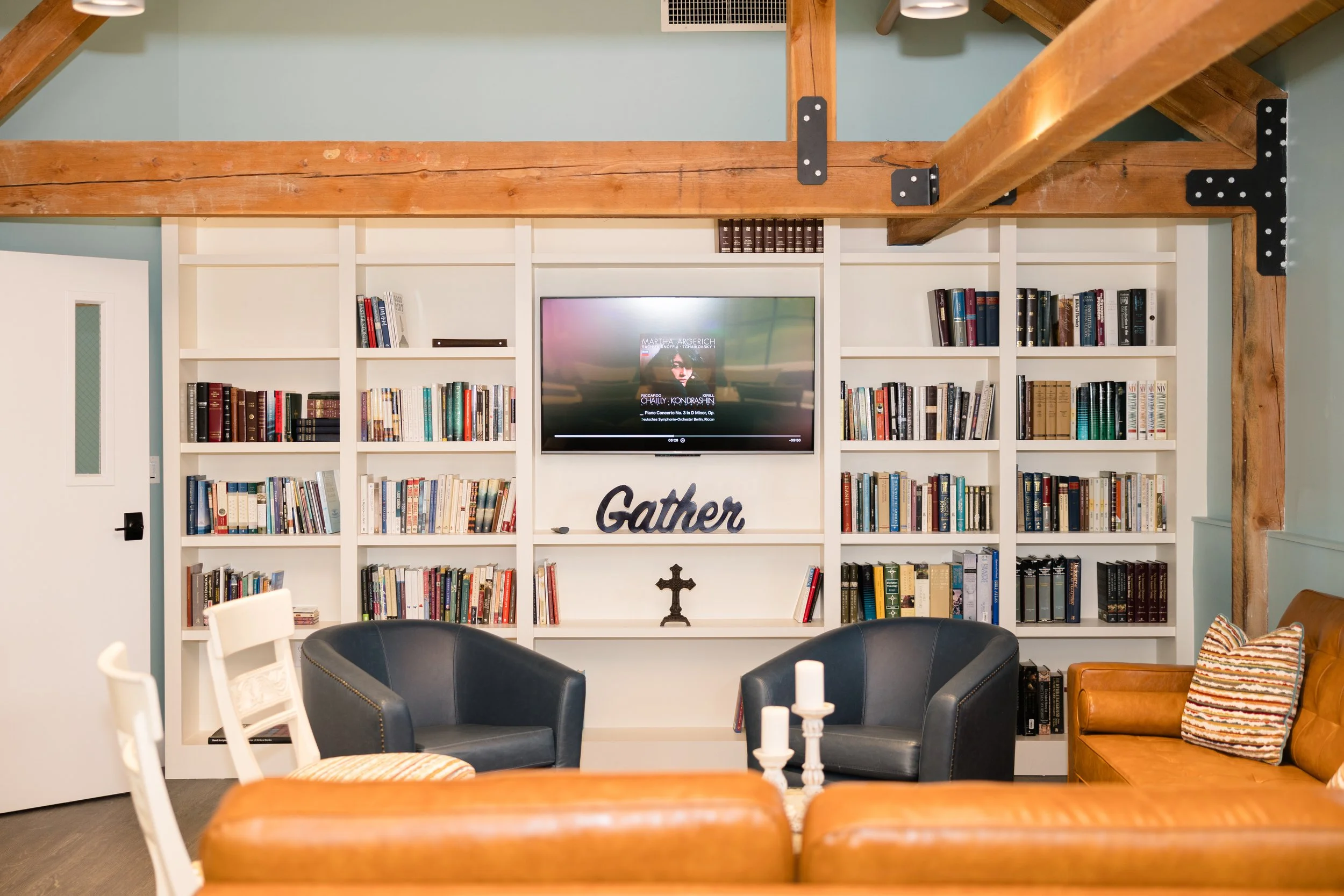 Living room with white built-in bookshelf filled with books, a mounted TV, a decorative sign that says 'Gather', a small cross figurine, a tan sofa with striped pillow, two black armchairs, and a white chair with a striped cushion. Exposed wooden bea