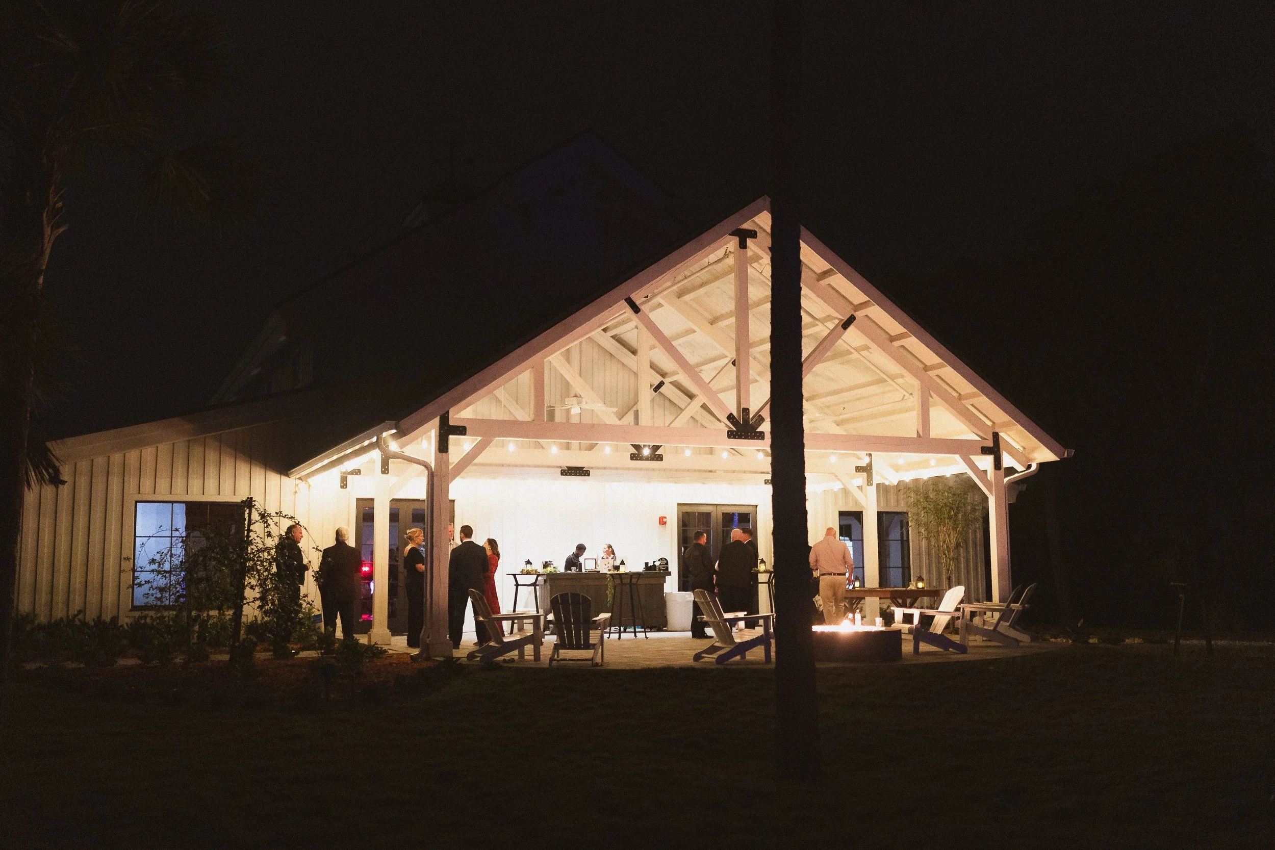 A nighttime outdoor gathering inside a lit pavilion with people conversing and a bar station, with patio chairs and a fire pit in front.