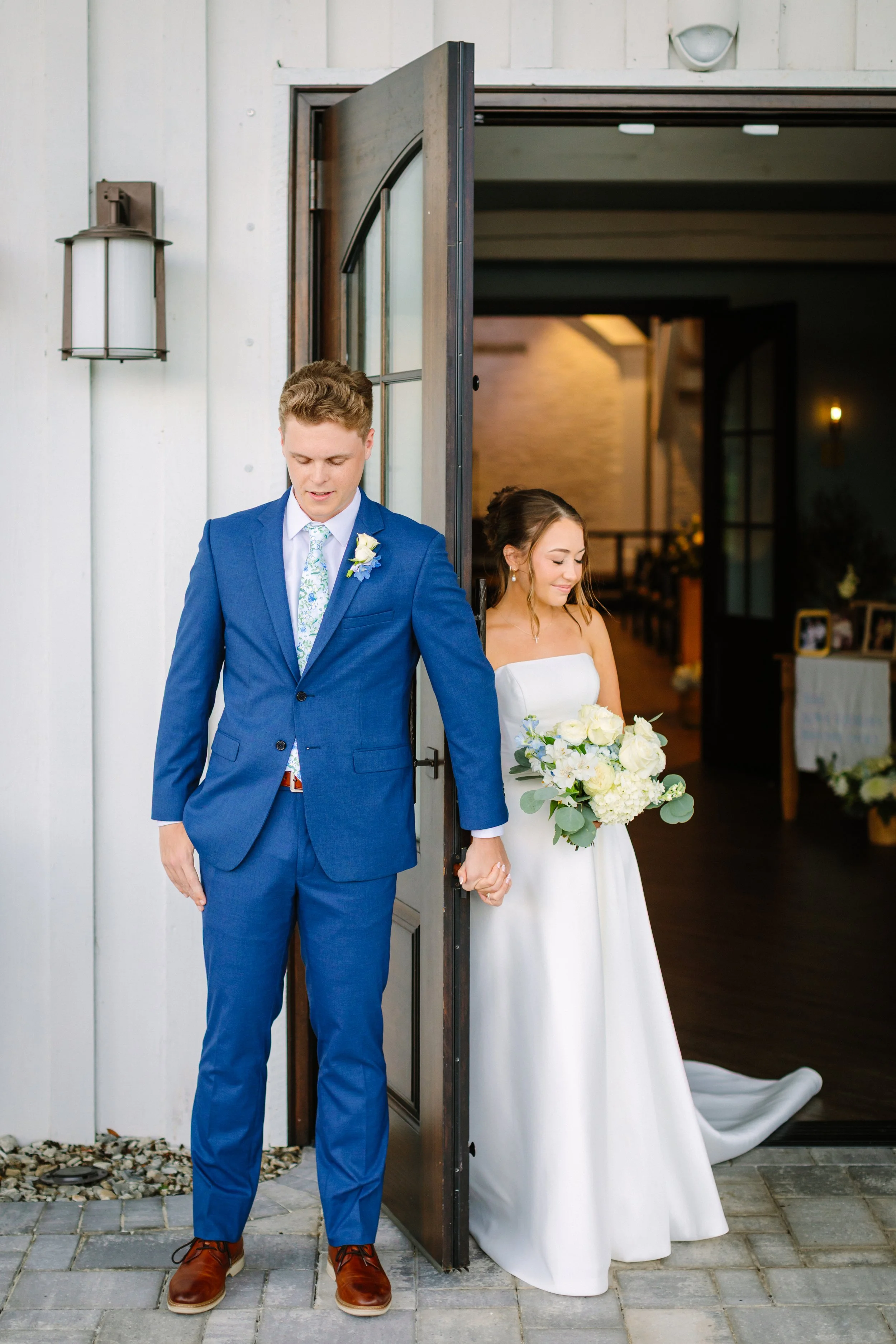 A bride and groom holding hands in a doorway, with the bride wearing a strapless white wedding dress and holding a bouquet of white flowers, while the groom wears a blue suit and tie.