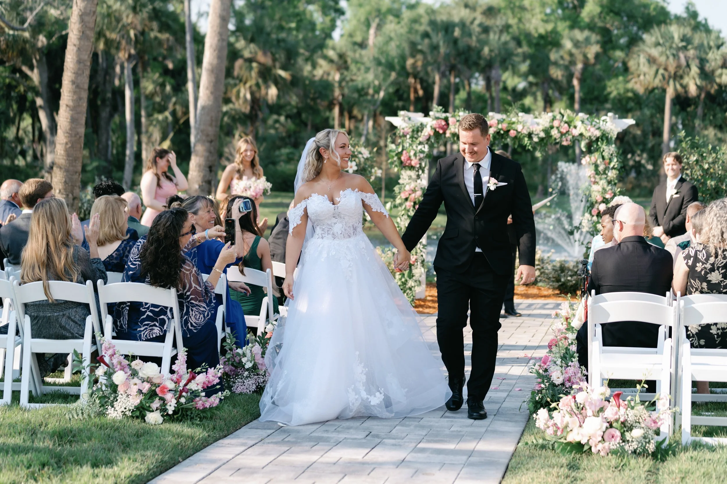 Bride and groom holding hands walking down the aisle at an outdoor wedding ceremony, surrounded by seated guests and floral decorations.