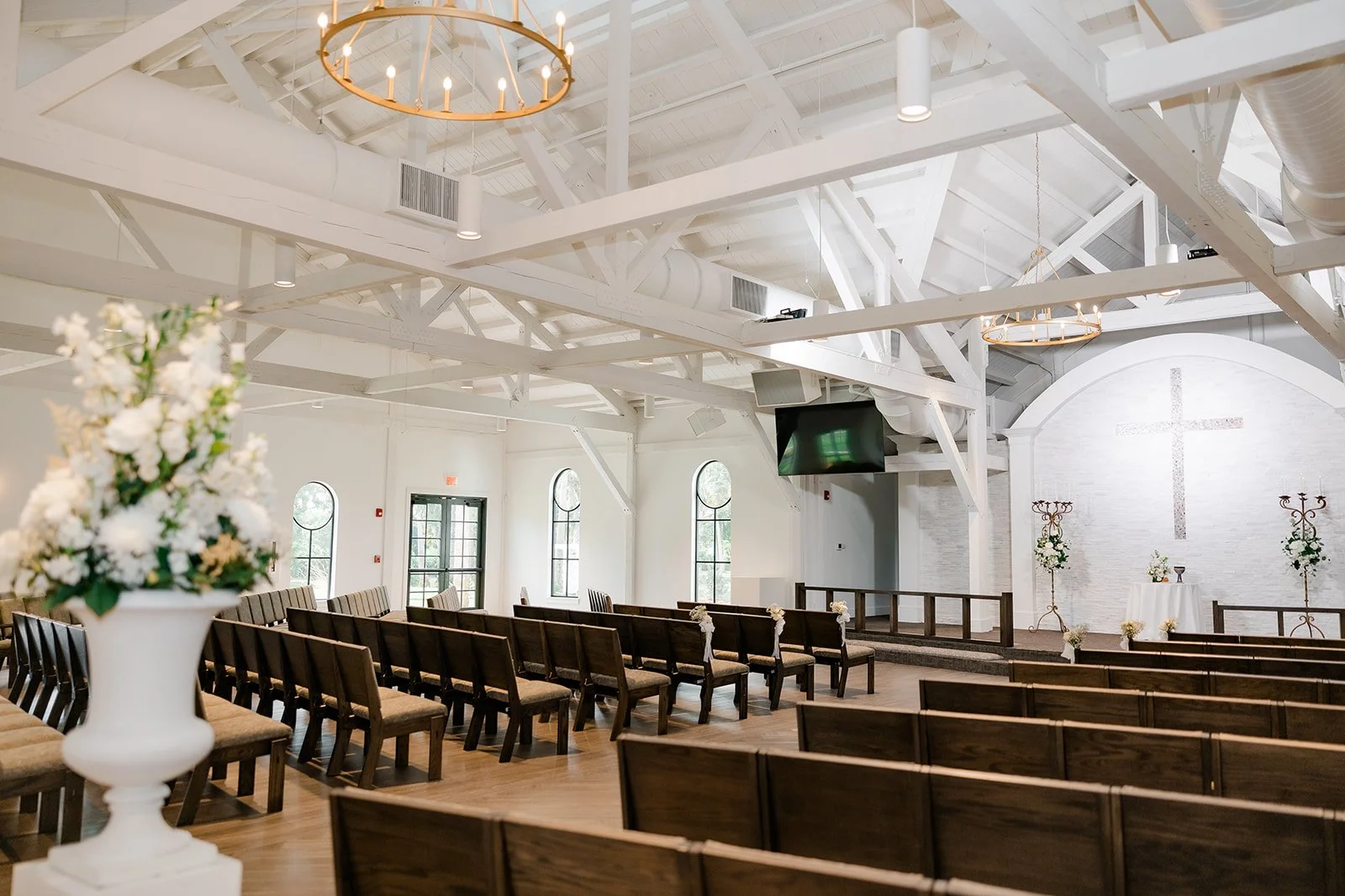 Empty wedding chapel with wooden pews, a white altar with floral arrangements, a cross on the wall, and large windows.