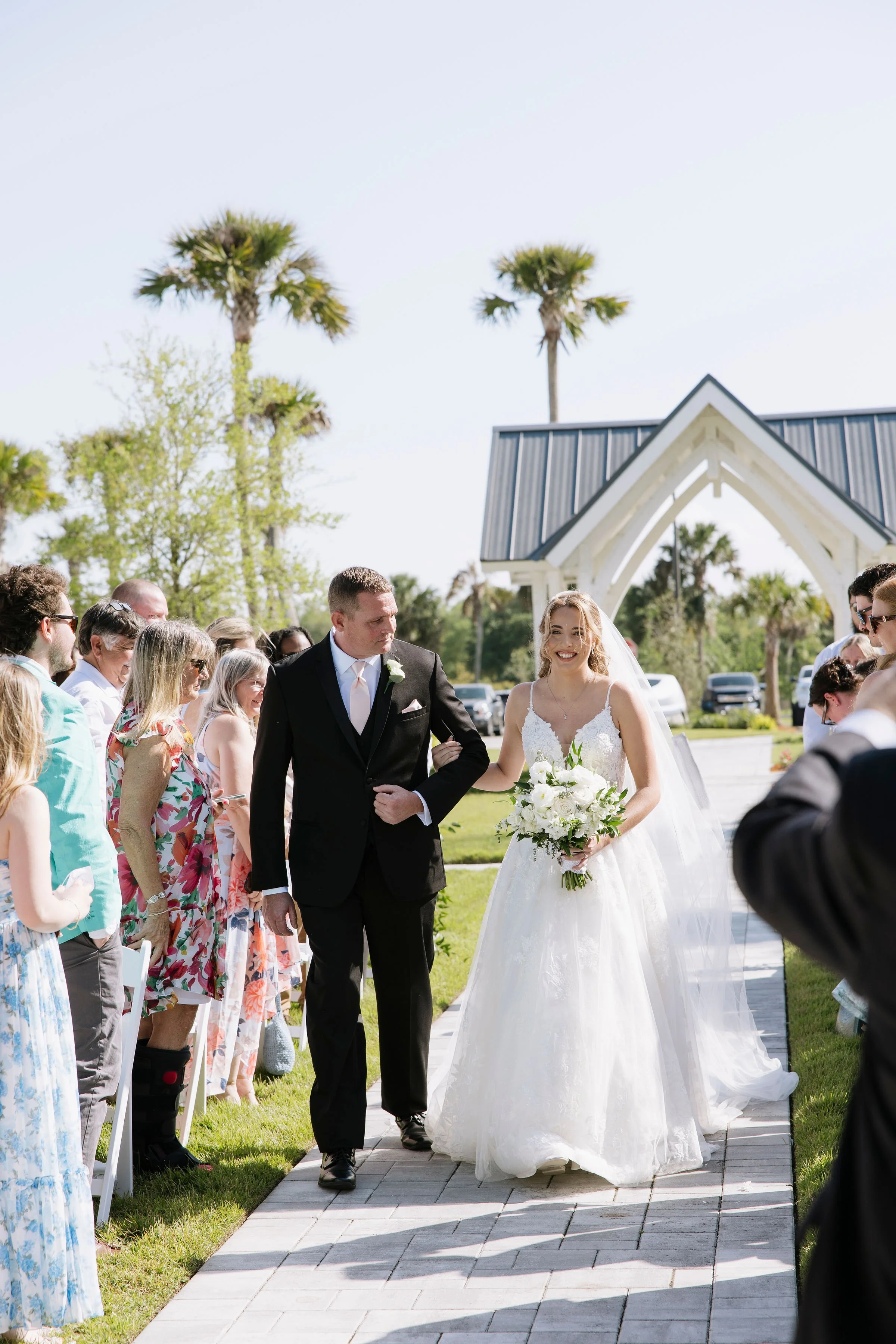 A bride in a white wedding gown with a bouquet, walking down the aisle with a man in a black suit, surrounded by seated wedding guests outdoors on a sunny day.