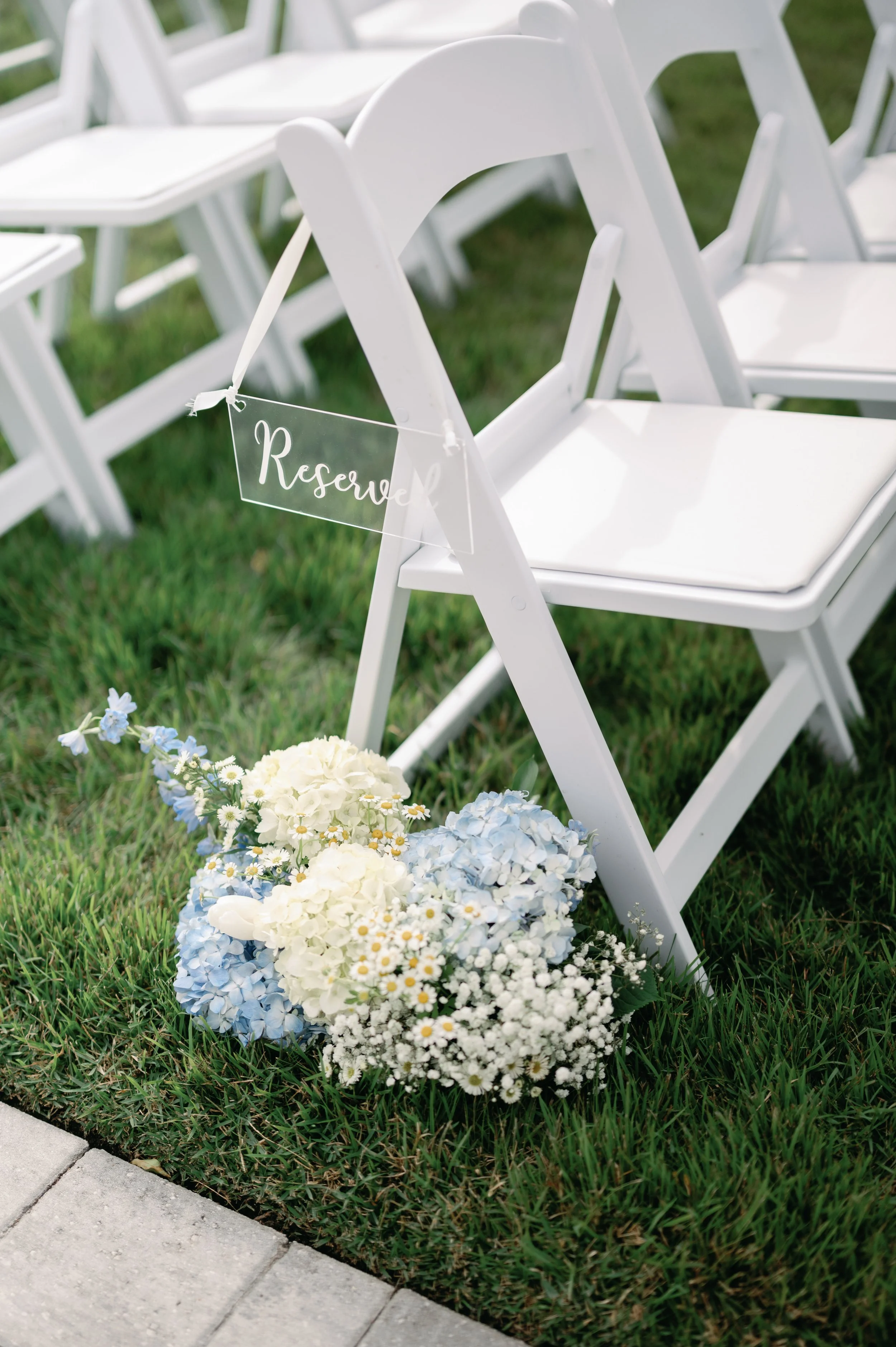 White chairs with a reserved sign, decorated with blue and white flowers, on a grassy area.