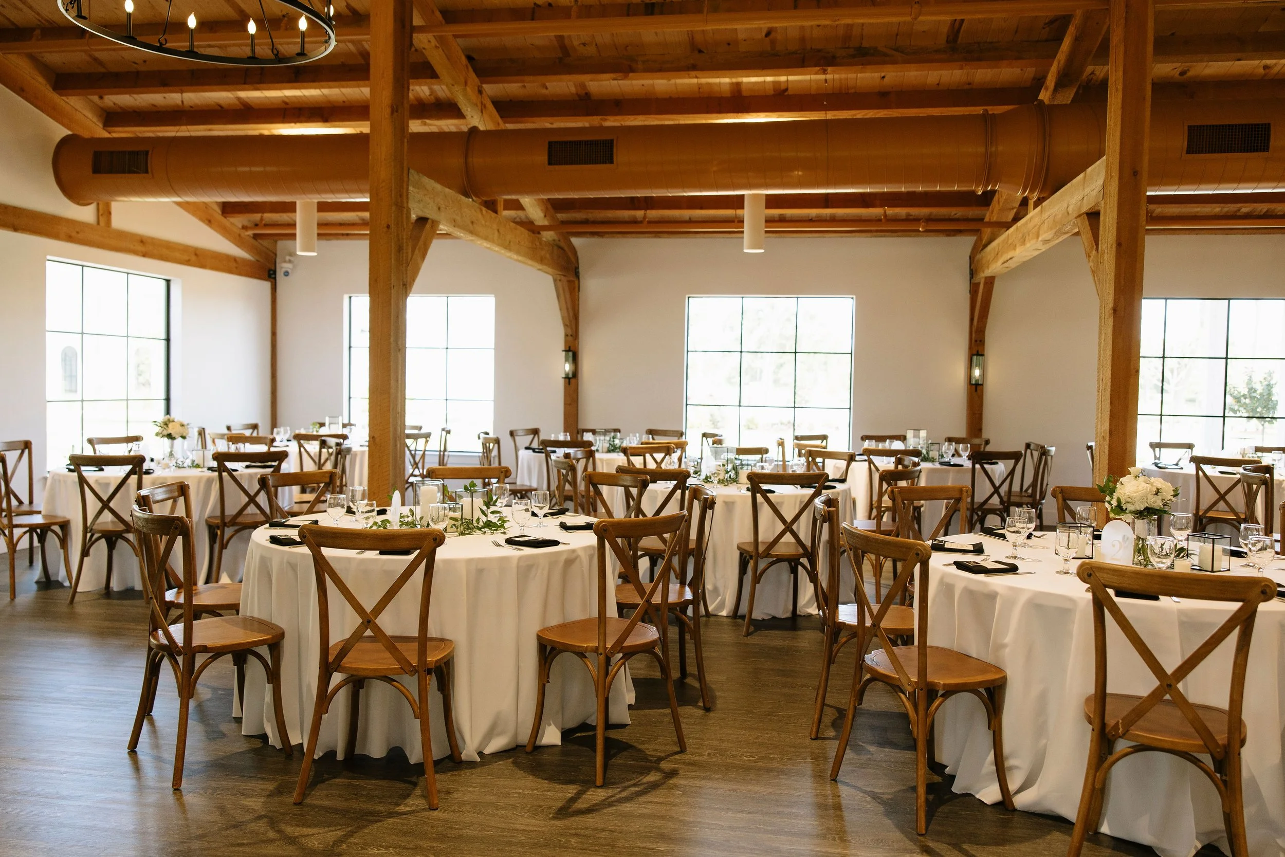 Elegant banquet hall with round tables covered in white tablecloths, wooden chairs, floral centerpieces, and large windows with natural light.