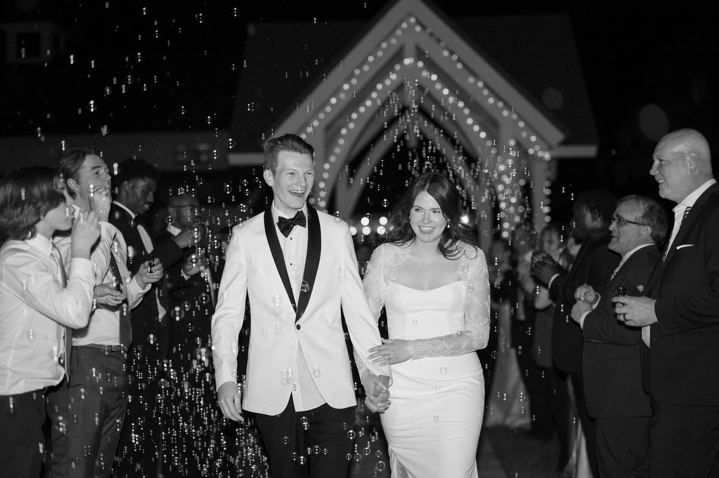 A newlywed couple walks hand-in-hand through a crowd of friends and family outside at night, with bubbles floating around, in a wedding celebration setting.