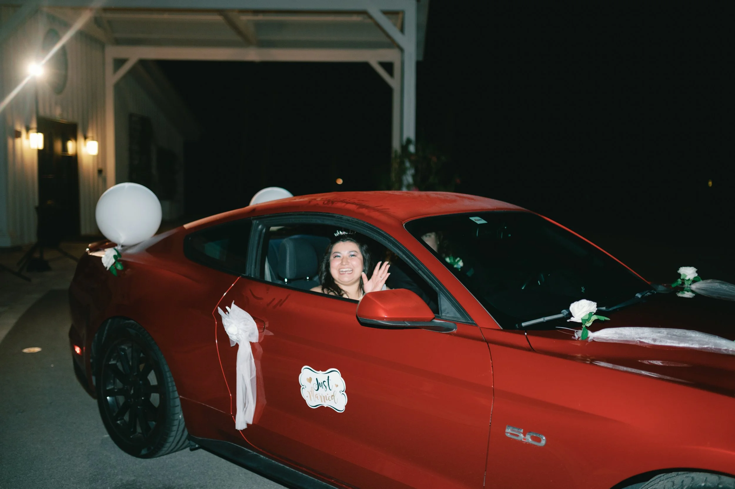 A woman sitting inside a red sports car, smiling and waving. The car is decorated with white balloons, flowers, and ribbons, indicating a celebration or wedding. A "Just Married" sign is attached to the car door. It is nighttime, outside a building w