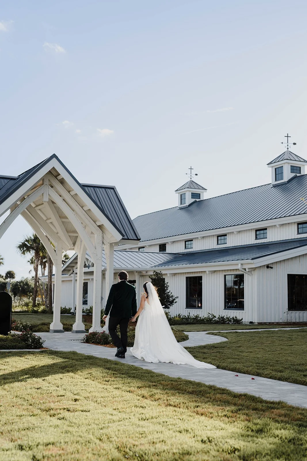 A bride and groom walking hand in hand outside a white church with a metal roof on a sunny day.